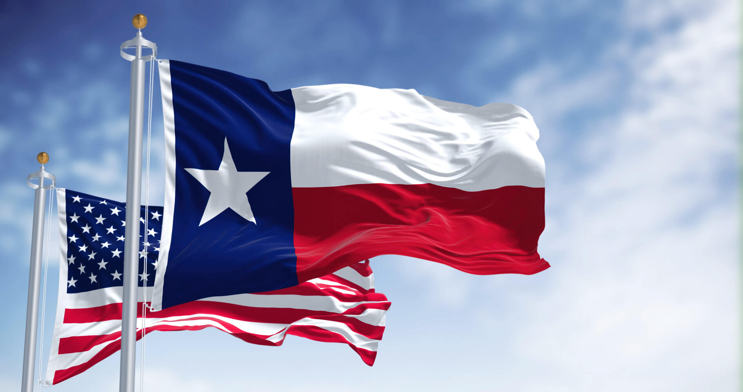 Two flags waving against a cloudy blue sky. The foreground flag is the Texas state flag, featuring a white star on a blue vertical stripe with horizontal white and red stripes. The background flag is the United States flag, showing stars and stripes.