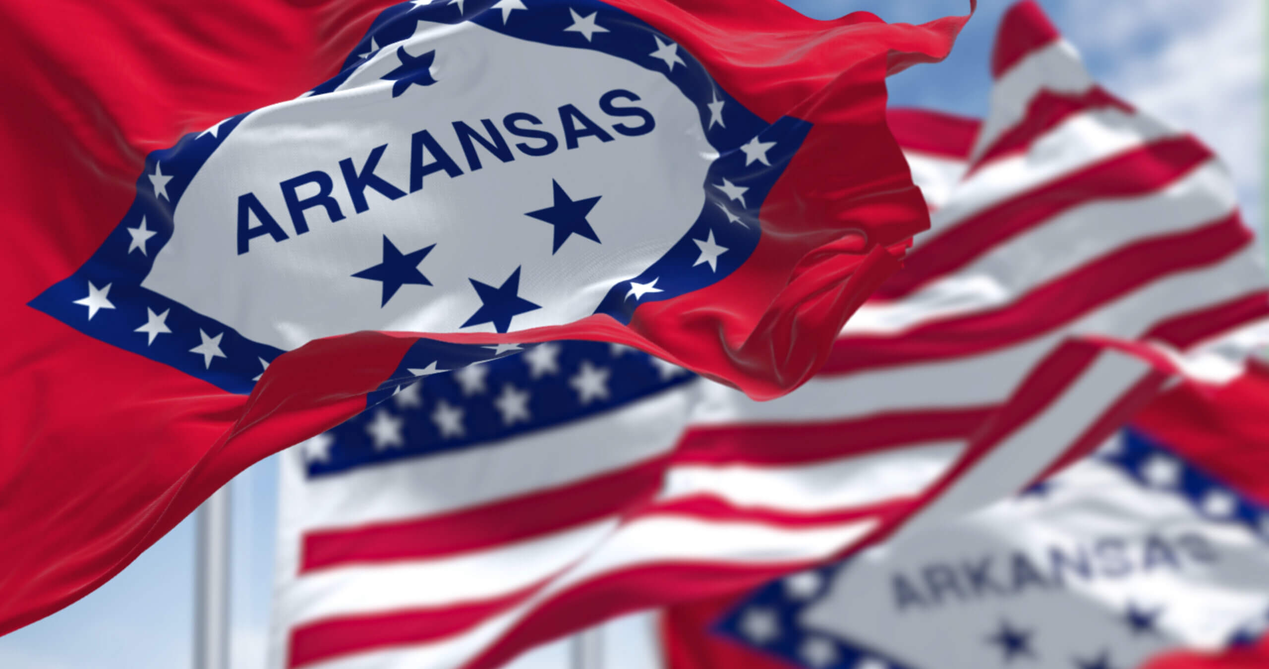 Arkansas state flags waving in the wind against a blue sky. The flags feature a red design with a white diamond at the center, surrounded by blue stars. The word "Arkansas" is prominently displayed in blue inside the diamond.