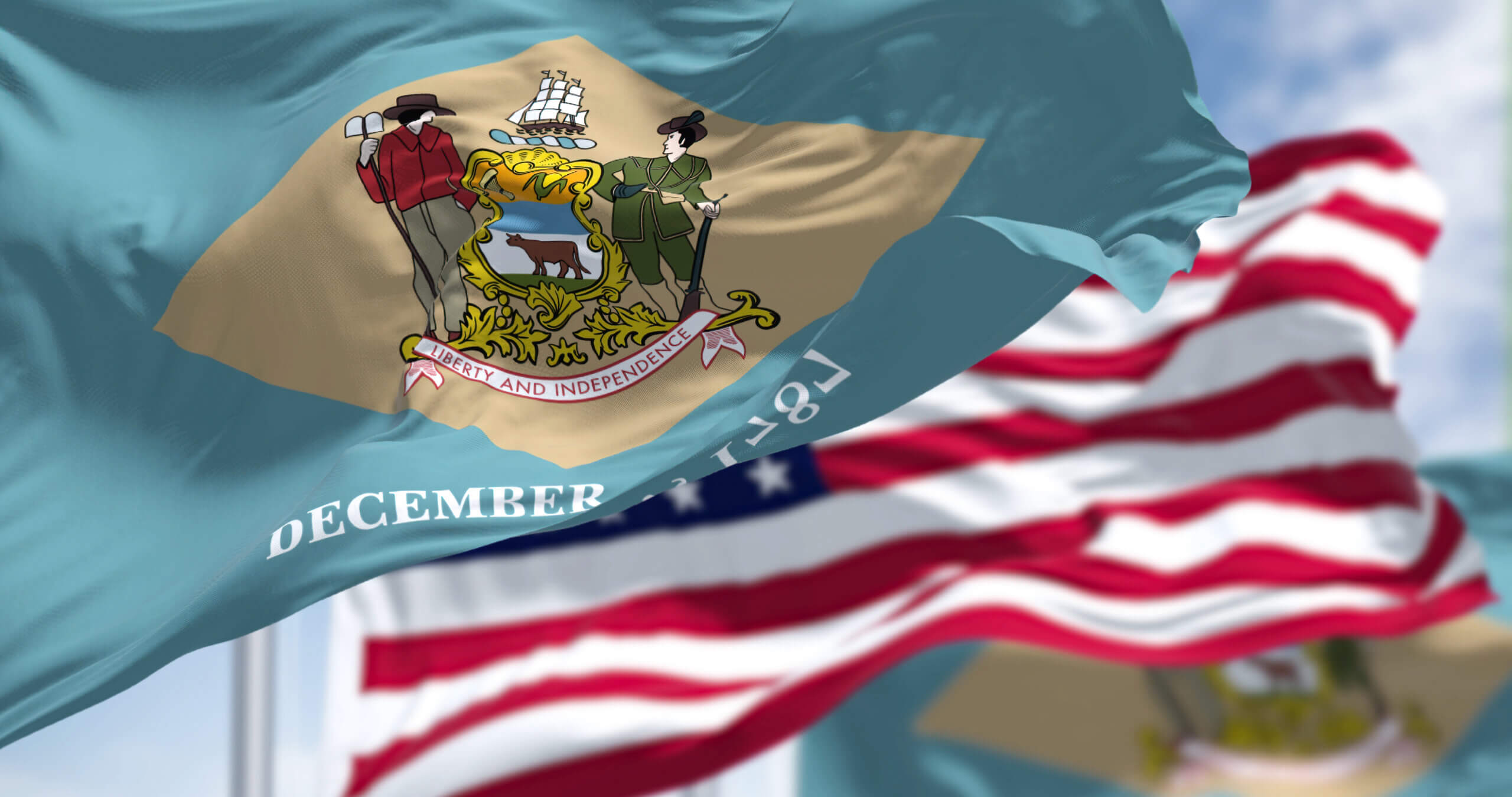 The image shows the Delaware state flag waving in the foreground, featuring the state coat of arms with a farmer, soldier, and ship above the motto "Liberty and Independence." In the background, the American flag is also visible.
