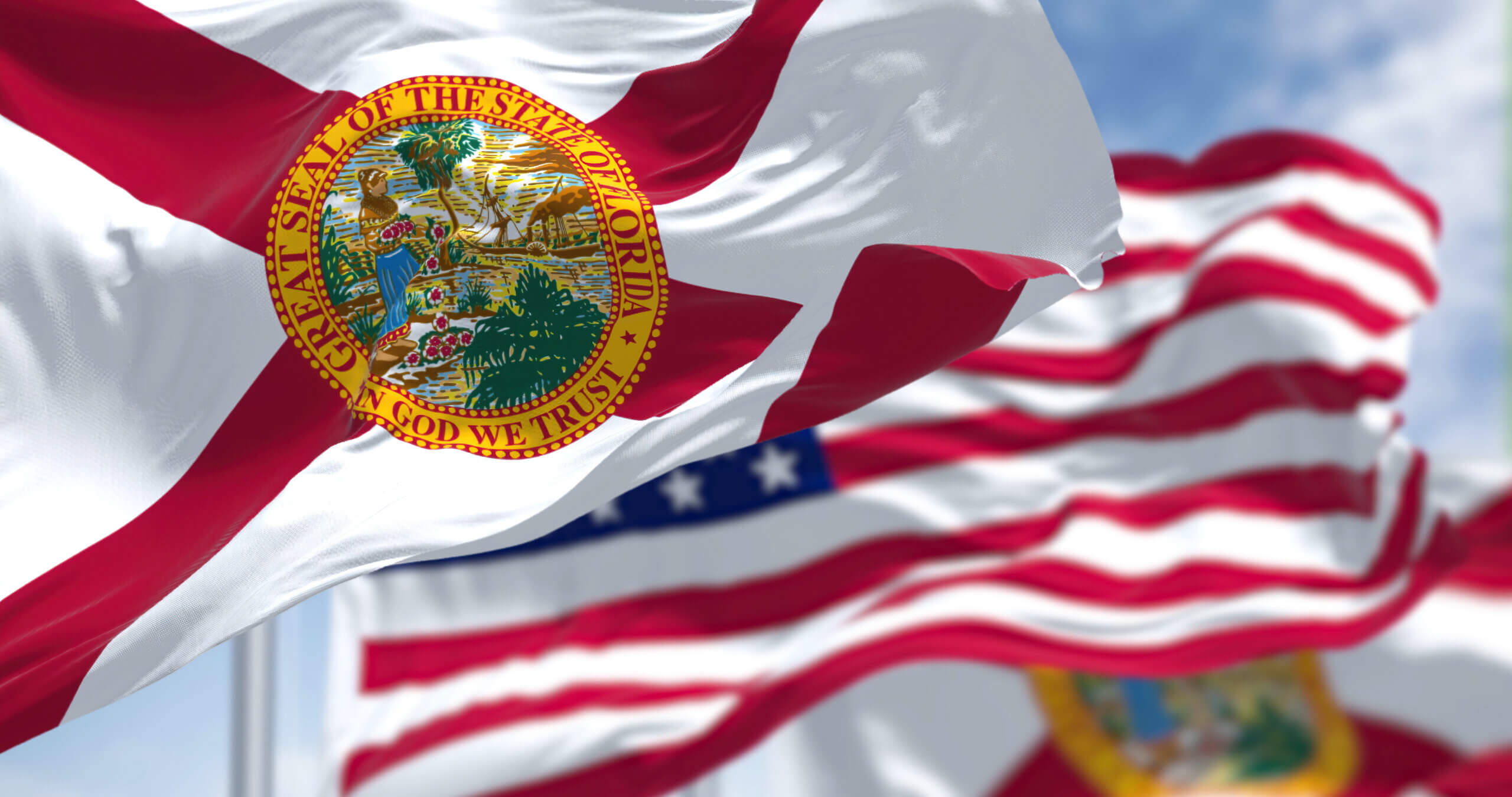 Florida state flag waving in the foreground with the seal depicting a Native American woman, a steamboat, and palm trees. In the background, multiple blurred American flags are waving against a blue sky.