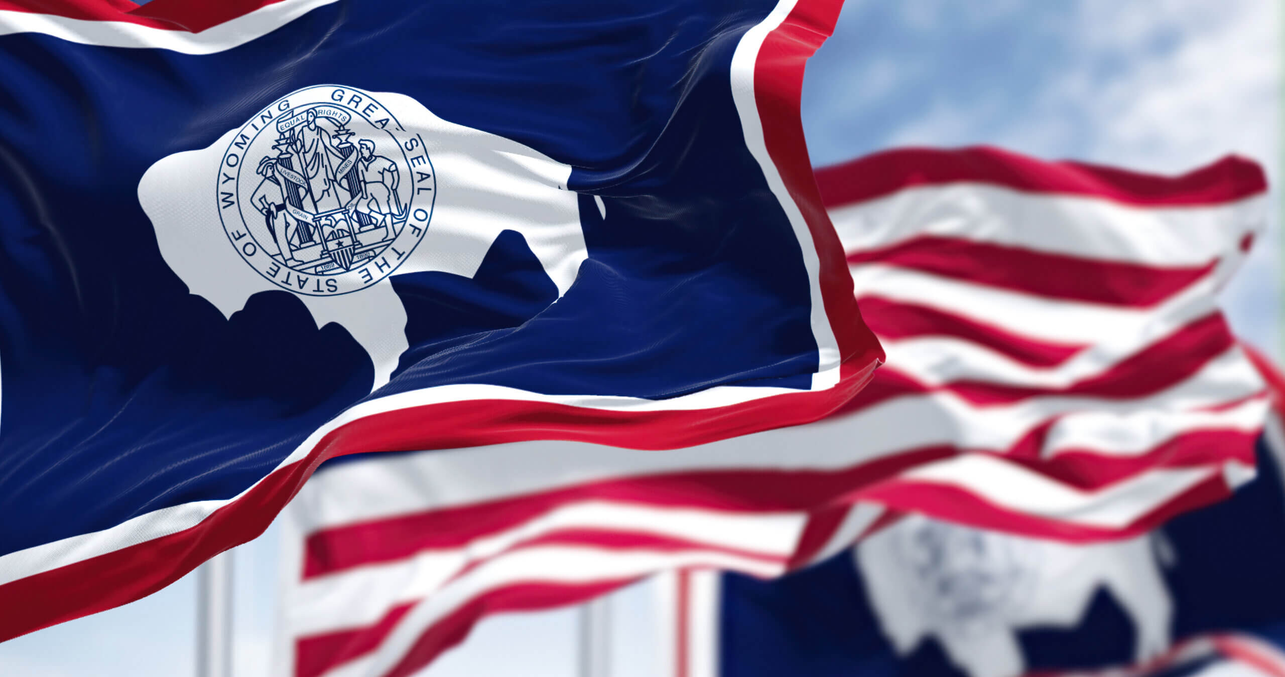 Flags featuring the Wyoming state design wave in the wind. The flag has a bison silhouette with the state seal in the center on a blue background, bordered by white and red stripes. The sky is partly cloudy.