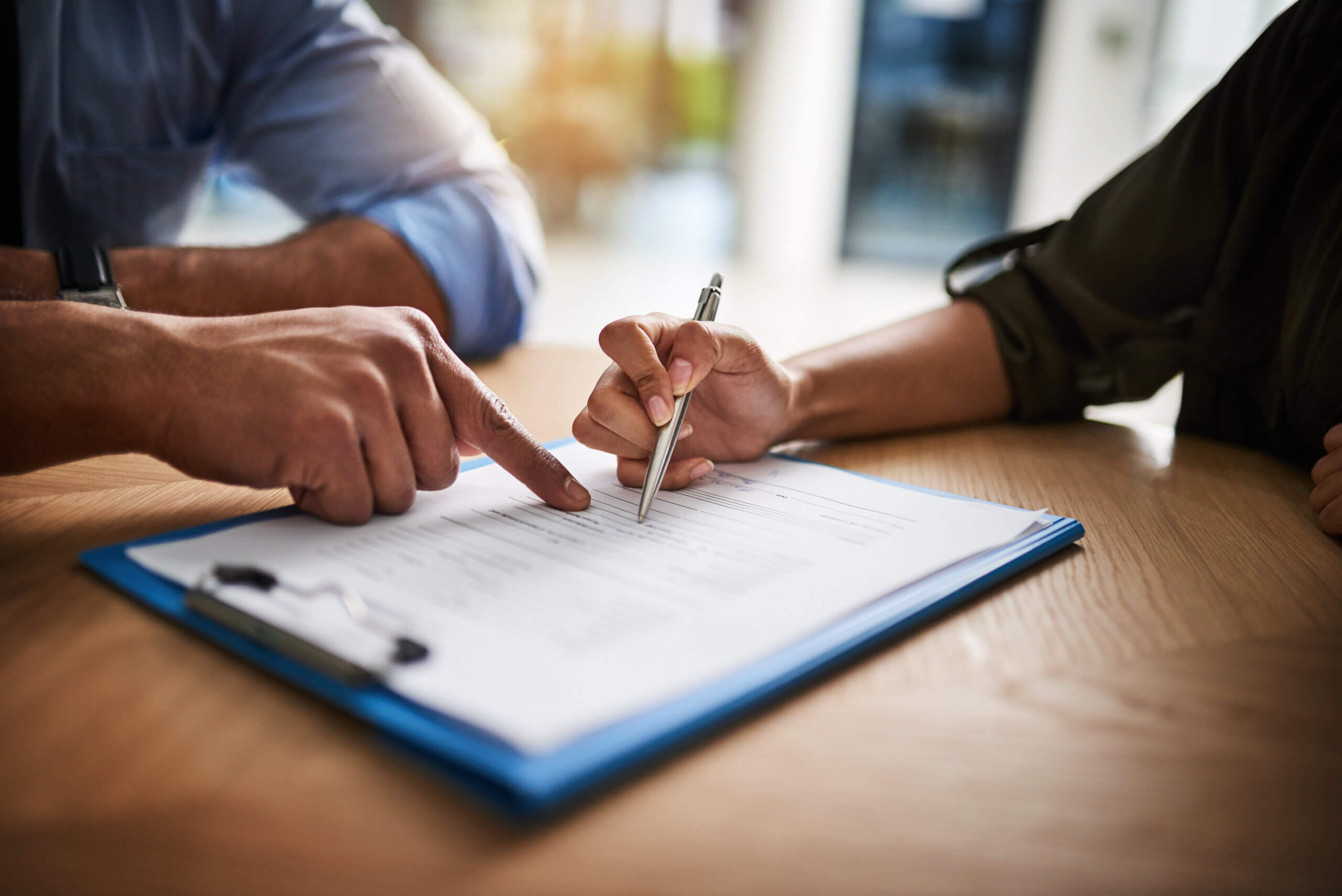 A person is filling out a form on a clipboard while another person points at the document. Both are seated at a wooden table, focusing on the paperwork. The scene conveys collaboration and attention to detail.