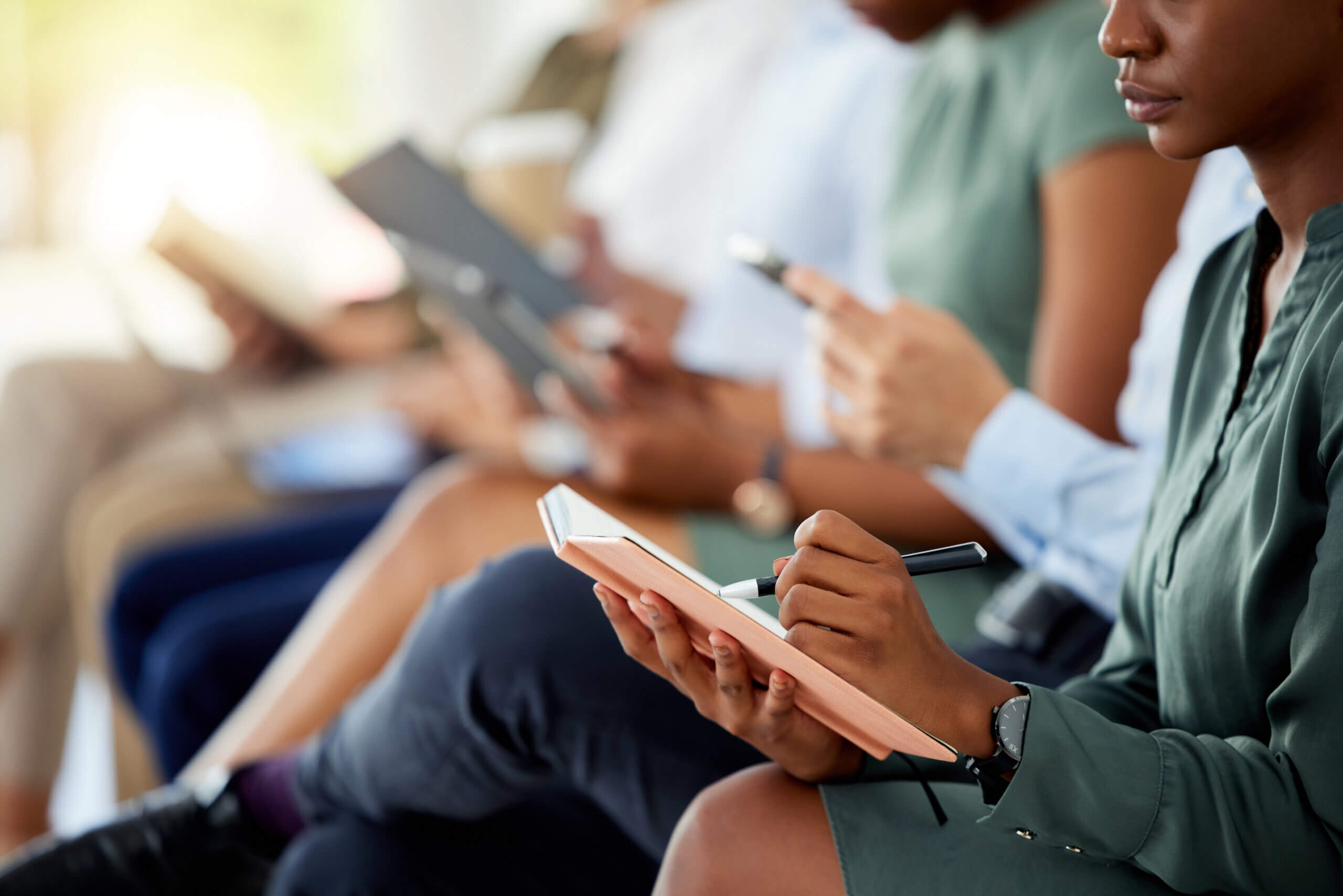 A row of people sitting and focusing on various tasks; one person in the foreground writes in a notebook, while others use smartphones and tablets. They appear to be attending a lecture or meeting, dressed in business casual attire.