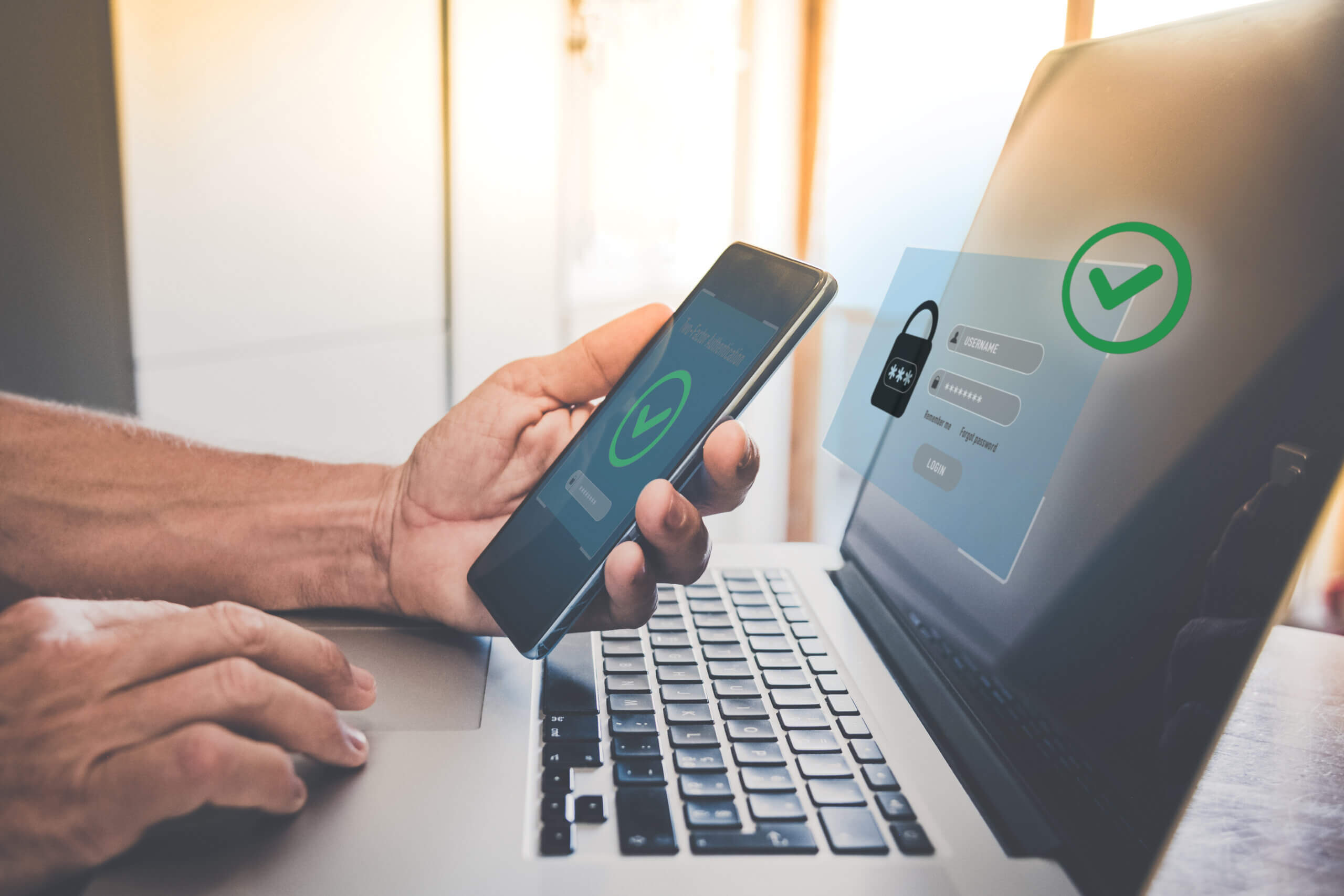 Person holding a smartphone with a green check mark on the screen, sitting in front of a laptop displaying a holographic padlock and check mark, symbolizing secure login and two-factor authentication. Sunlight filters in from the background.