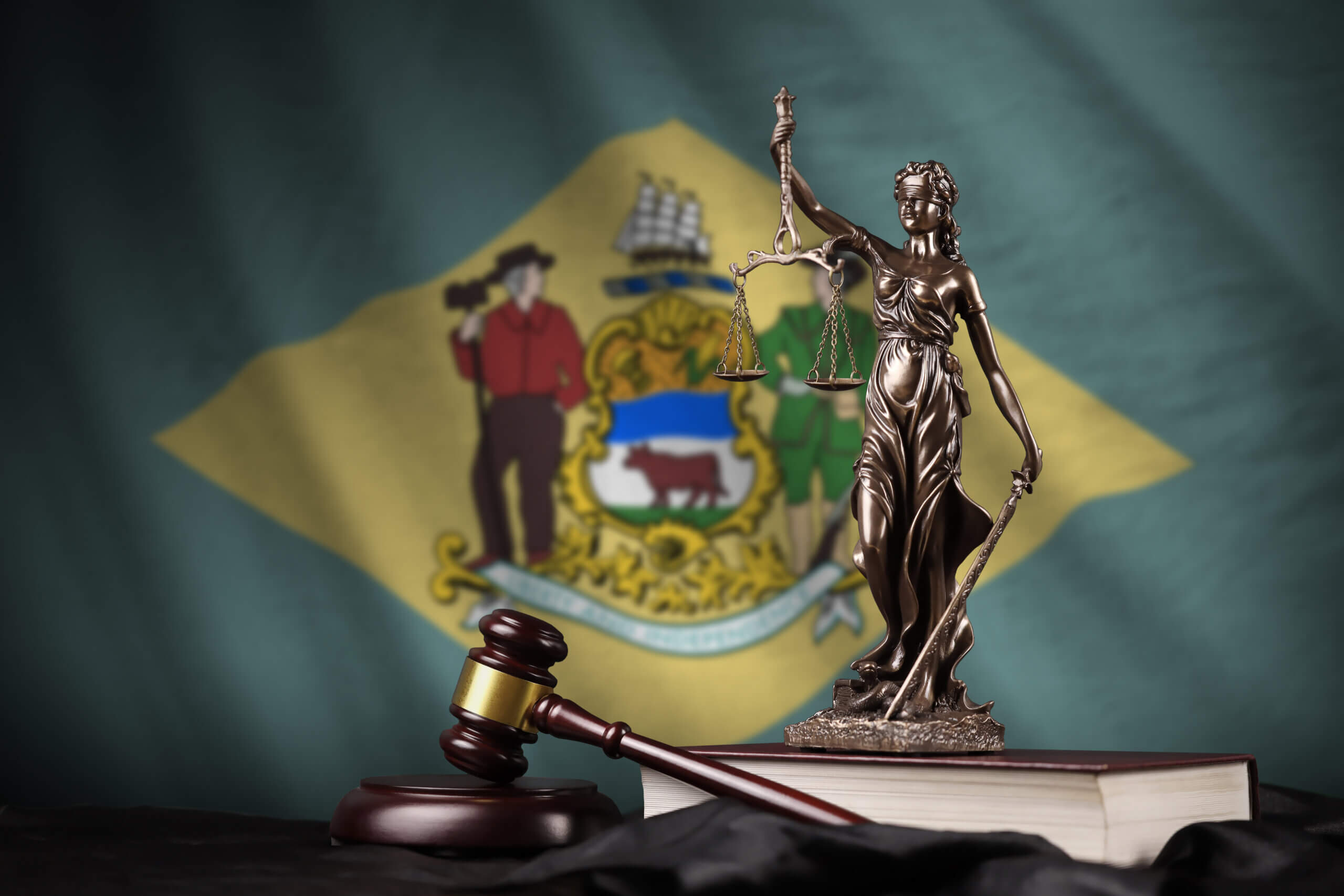 A bronze Lady Justice statue holds scales and a sword, standing on law books. A gavel lies nearby. The backdrop is the Delaware state flag, featuring a diamond shape with state symbols and figures on it.
