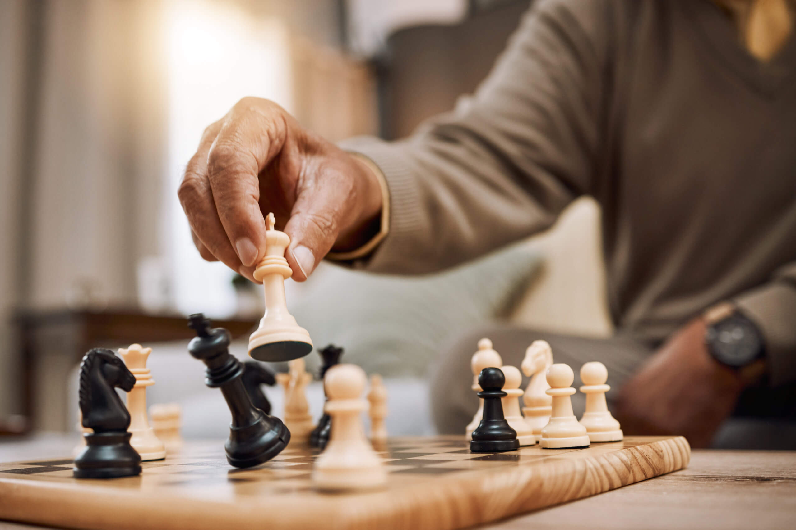 Close-up of a person's hand holding a white chess king piece above a chessboard. Various black and white pieces are in play. The player is wearing a brown sweater and a watch, and the scene is indoors with soft lighting.