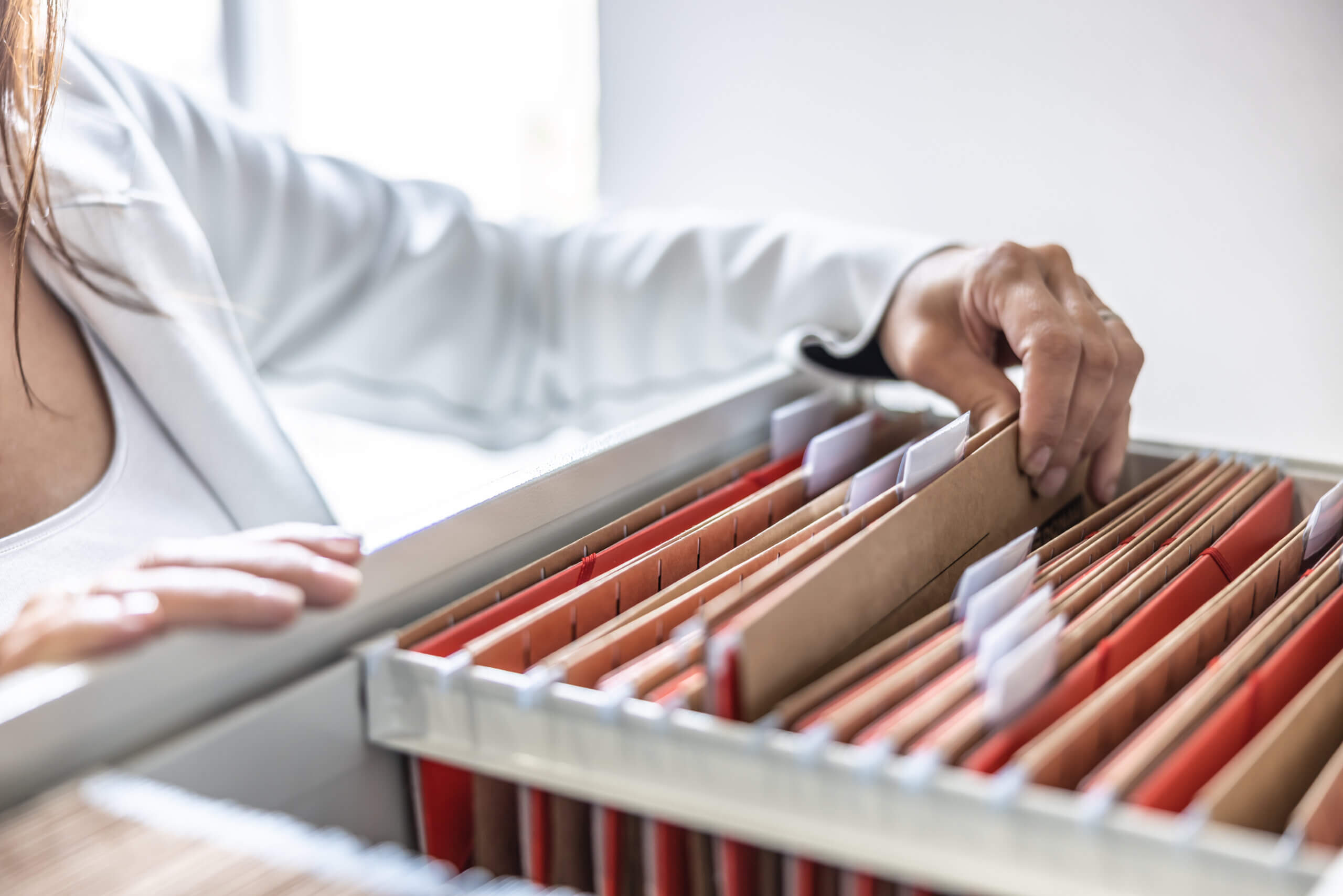 A person in a white jacket is organizing files in a filing cabinet. Their hand is sorting through labeled folders, which are neatly arranged in the drawer. The setting appears to be an office environment.
