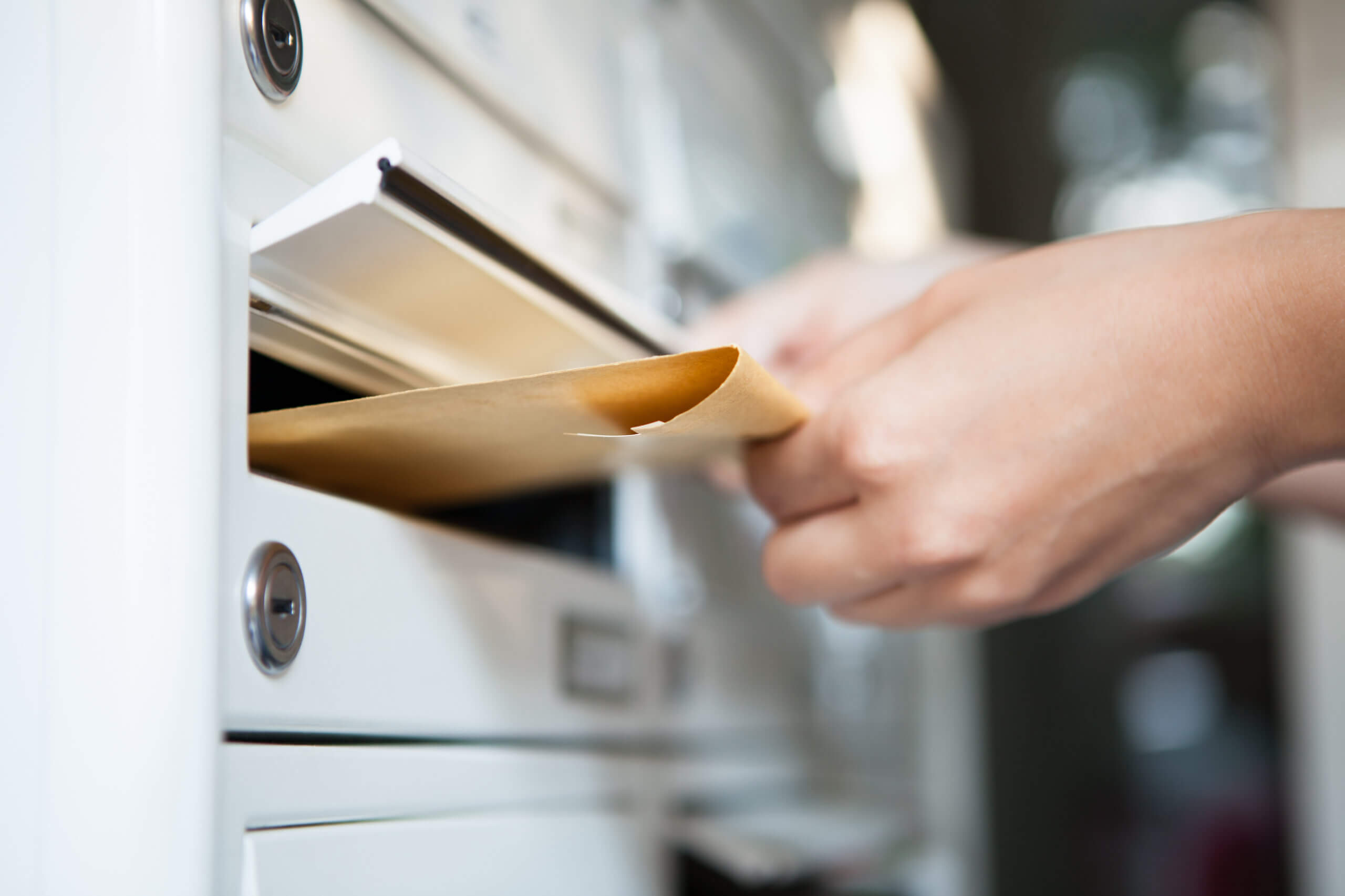Close-up of a hand inserting a brown envelope into a white mail slot. The mail slot is part of a larger panel with multiple horizontal slots, indicating a shared mailbox or apartment mail system.