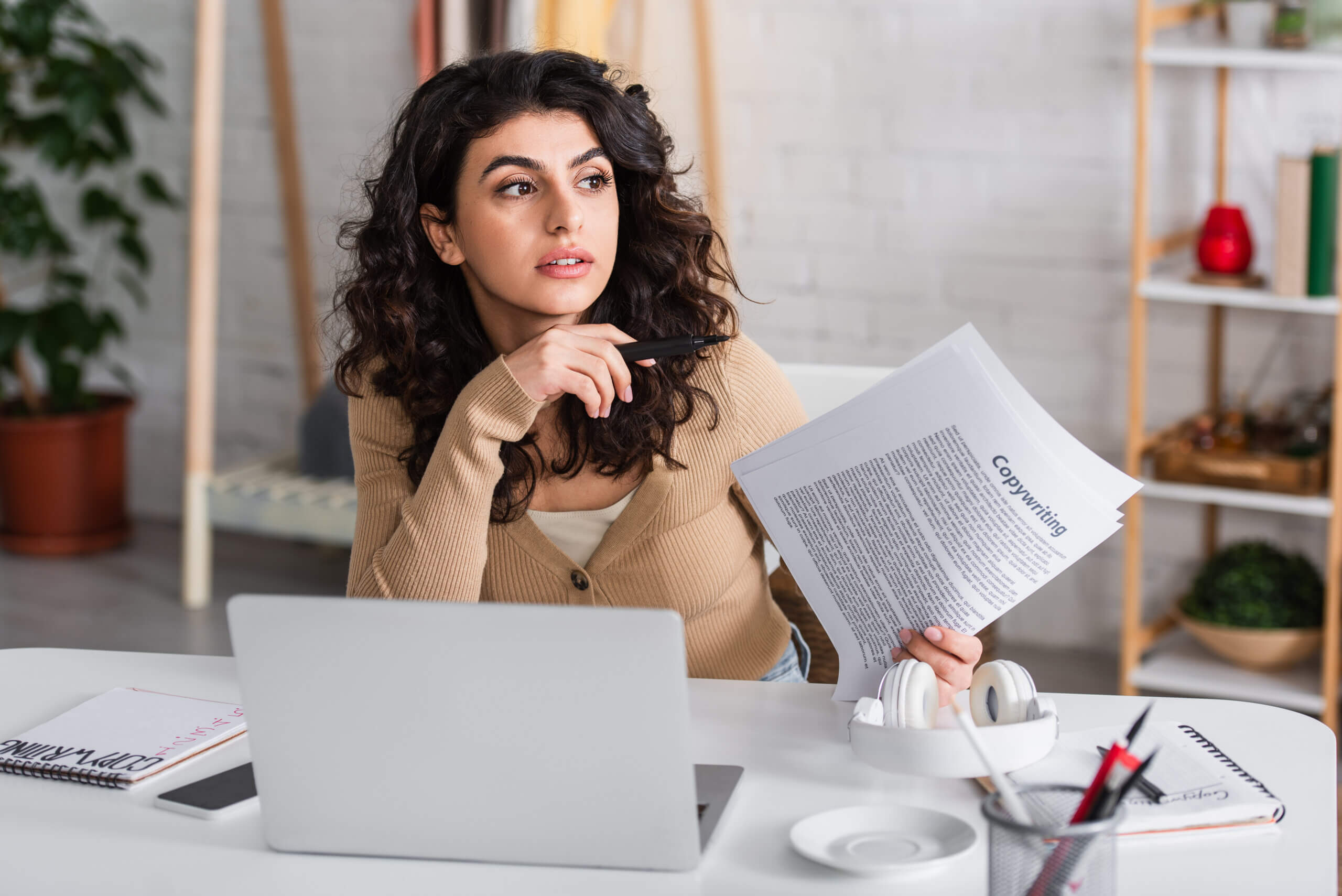 A woman with curly hair sits at a desk with a laptop, holding a pen and a document titled "Copyright." She appears thoughtful, with a notepad and headphones nearby. A plant and shelves are in the background.