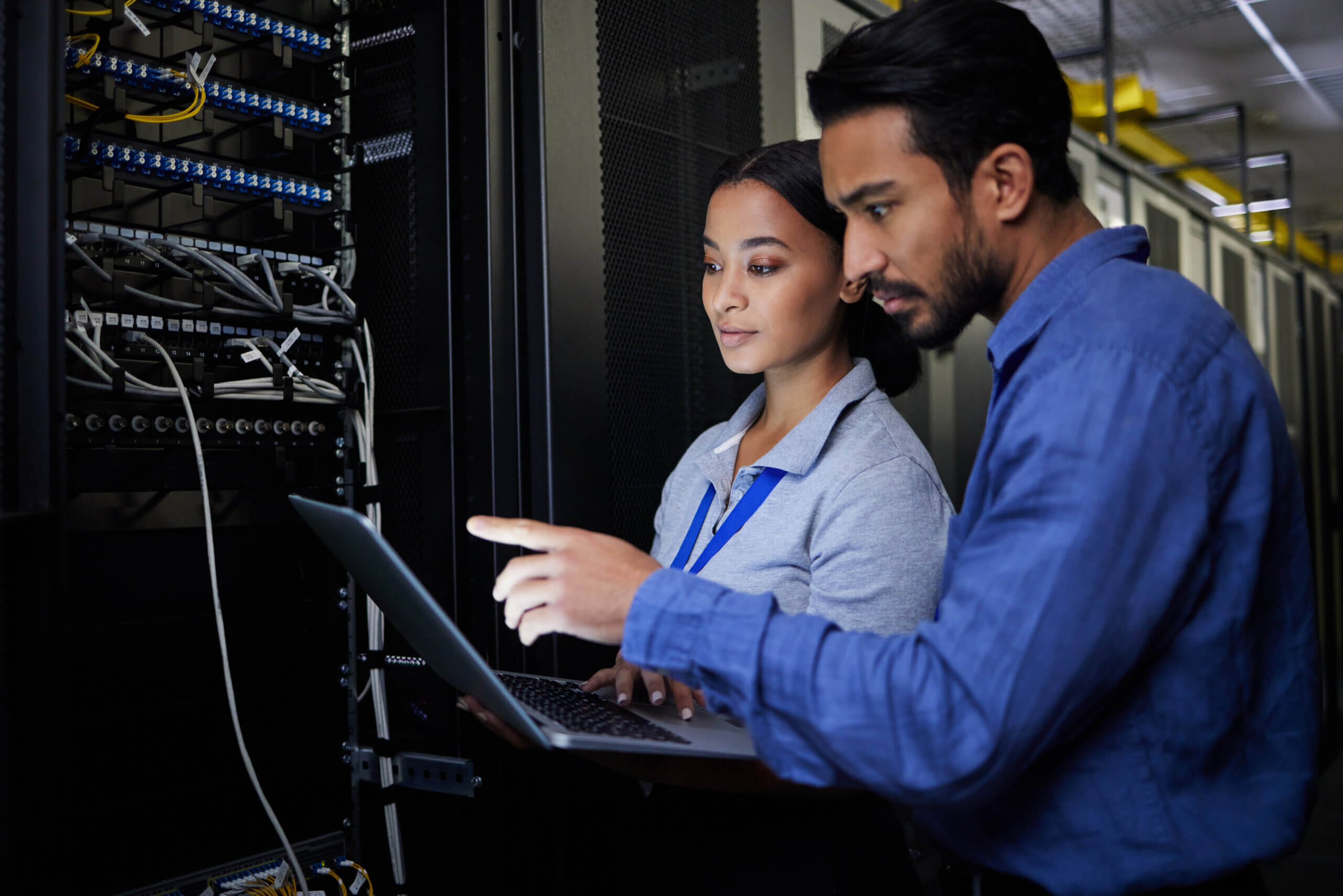 Two individuals in a server room examine data on a laptop. The woman holds the laptop, and the man gestures toward the screen. They are surrounded by server equipment, indicating a tech or IT environment.