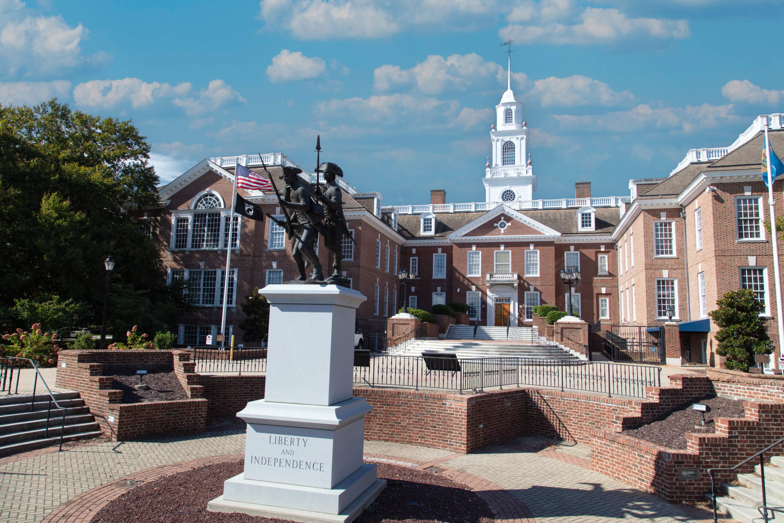 Statue of two soldiers on a pedestal labeled "Liberty and Independence" in front of a large brick building with white accents. The building has a clock tower and flag flying. The sky is clear with some clouds.