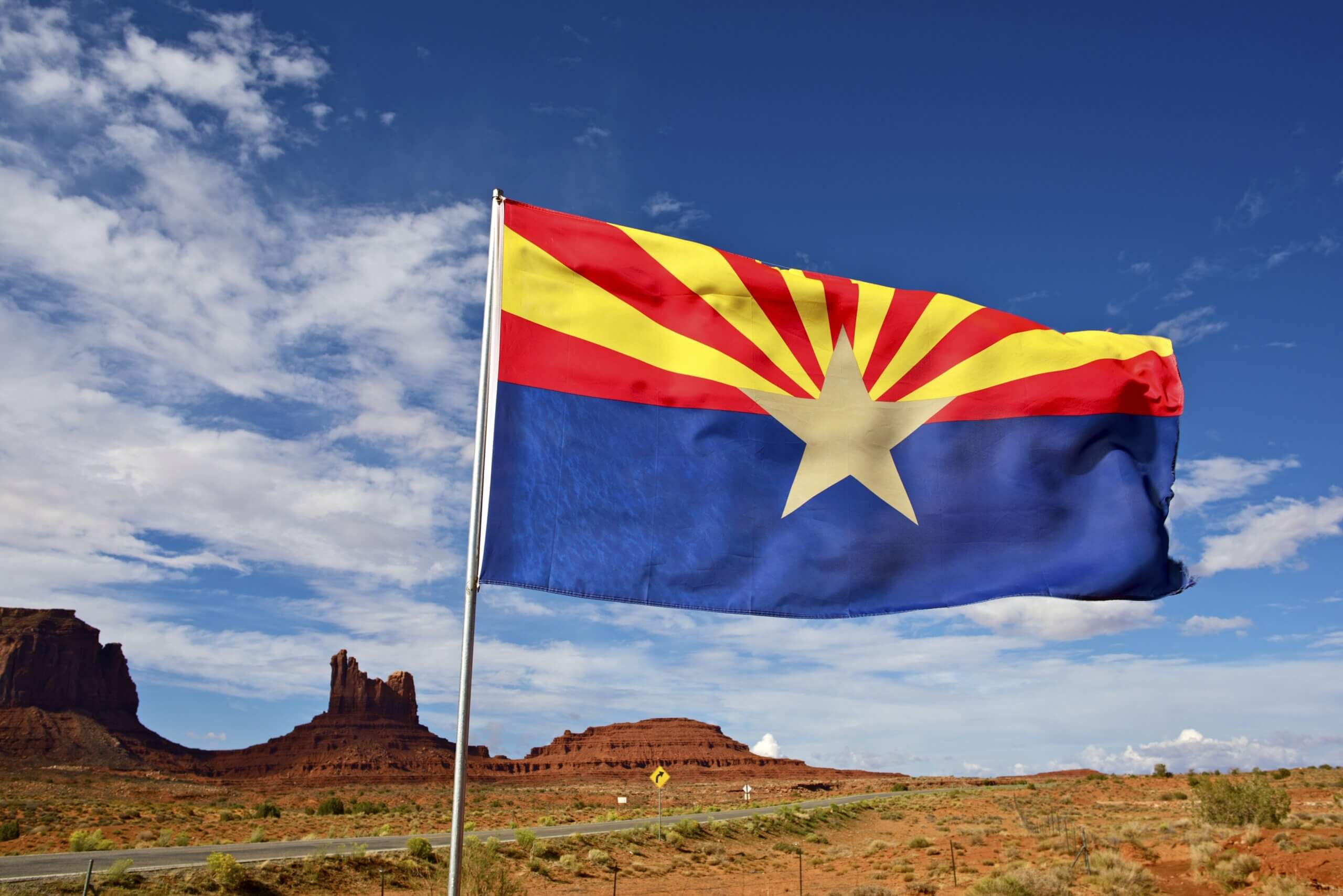 The Arizona state flag waving prominently with a backdrop of a vast desert landscape, featuring striking rock formations under a partly cloudy sky.