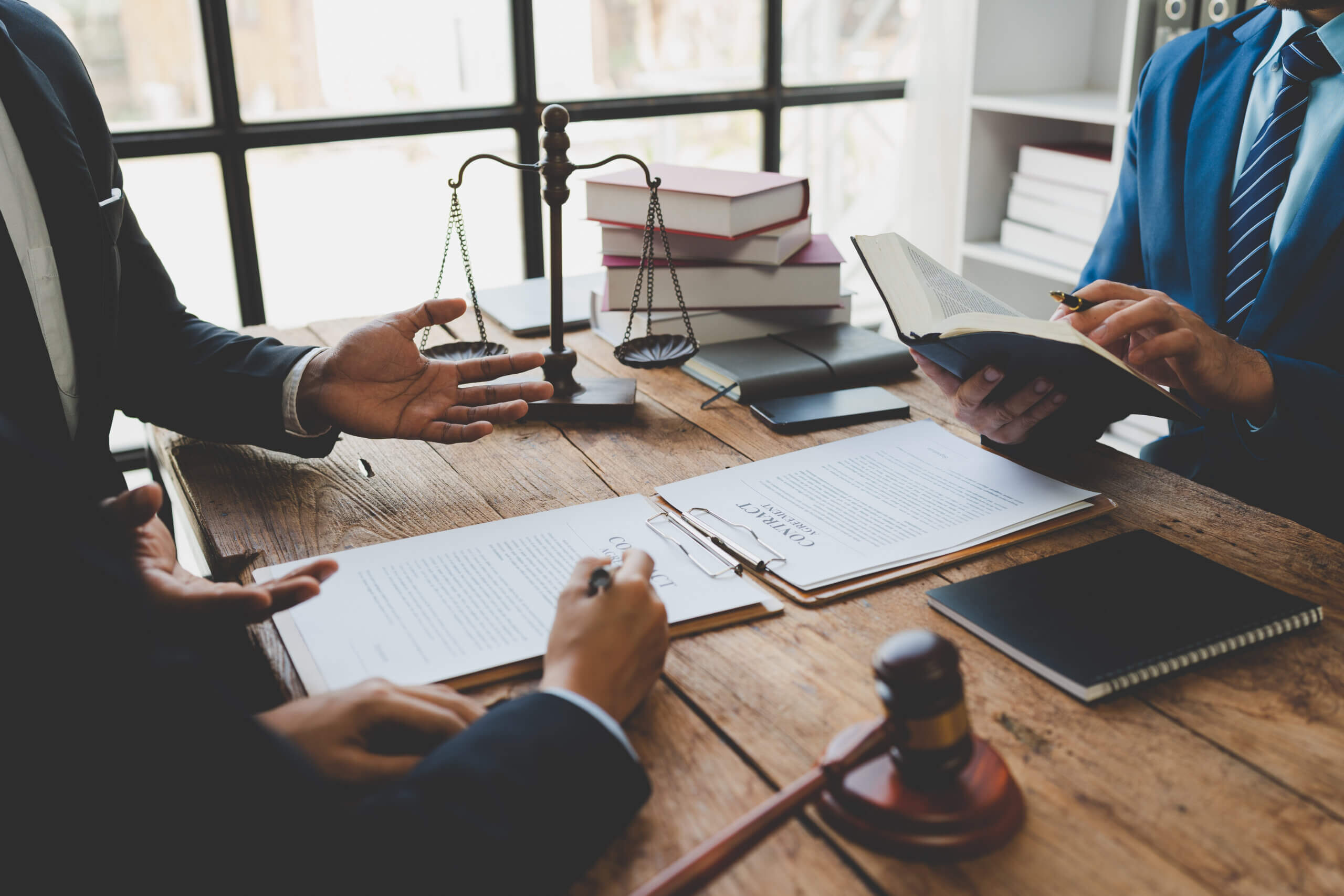 People in professional attire sit around a wooden table with legal documents, a gavel, scales of justice, and stacks of books. One person holds an open book while another gestures with their hand. Sunlight streams through a window in the background.
