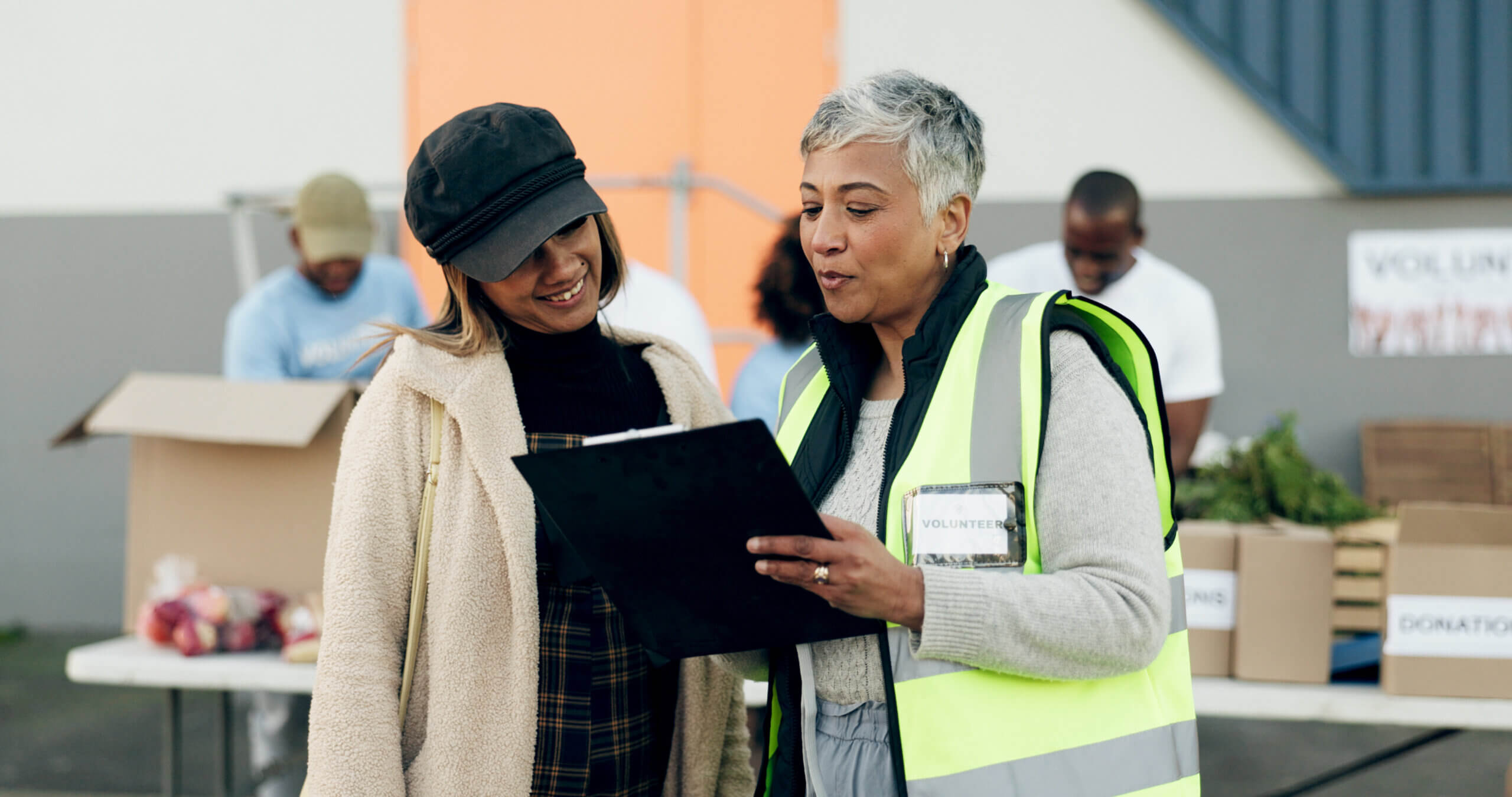 Two women, one in a yellow safety vest and holding a clipboard, stand together smiling. They are outdoors with volunteers organizing items on tables in the background. One woman wears a hat and coat, while the other has short gray hair.