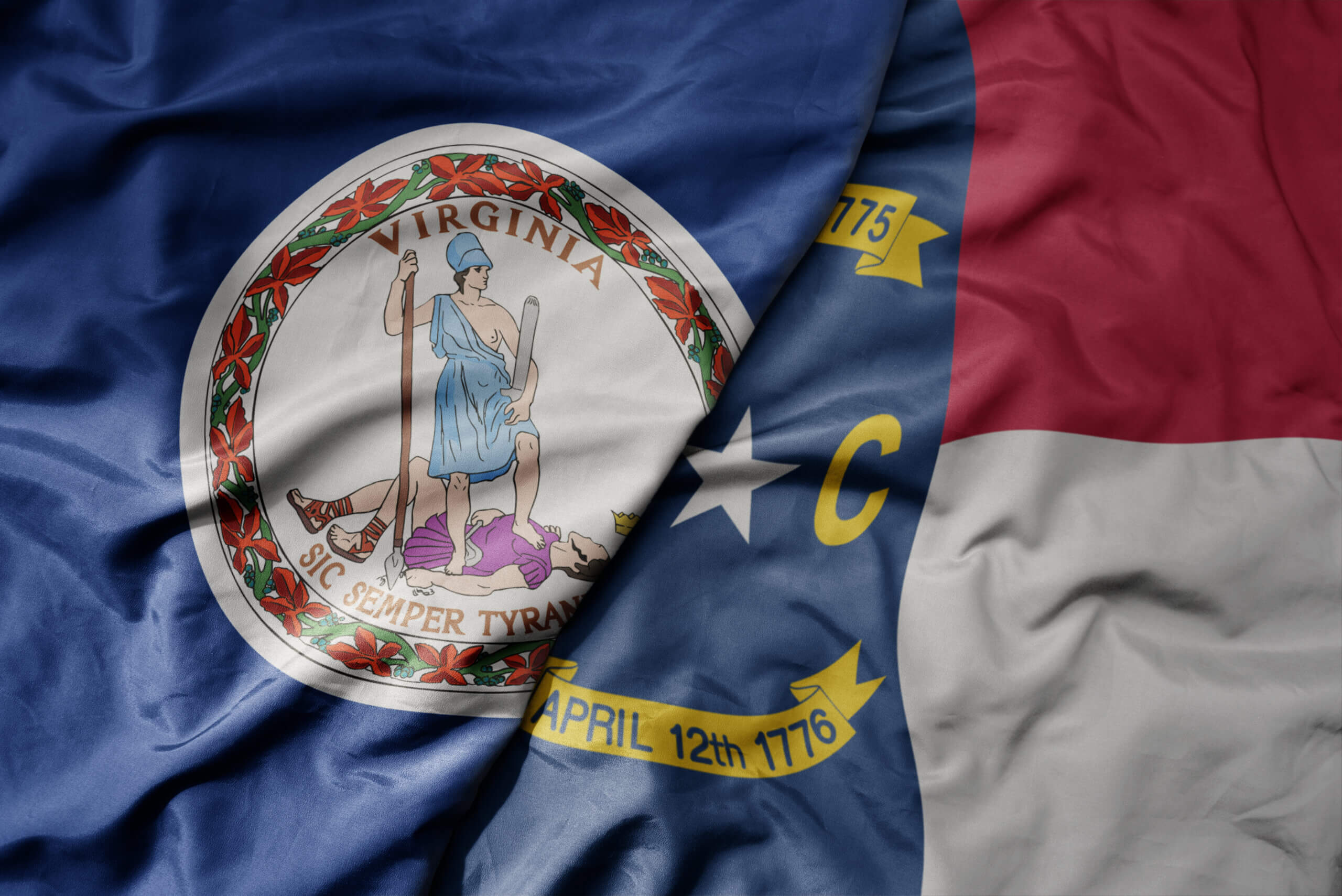 Two flags are draped side by side. The Virginia state flag with a seal featuring a figure and "Sic Semper Tyrannis," and the North Carolina state flag with a star, the letters "N" and "C," and the date "April 12th, 1776.