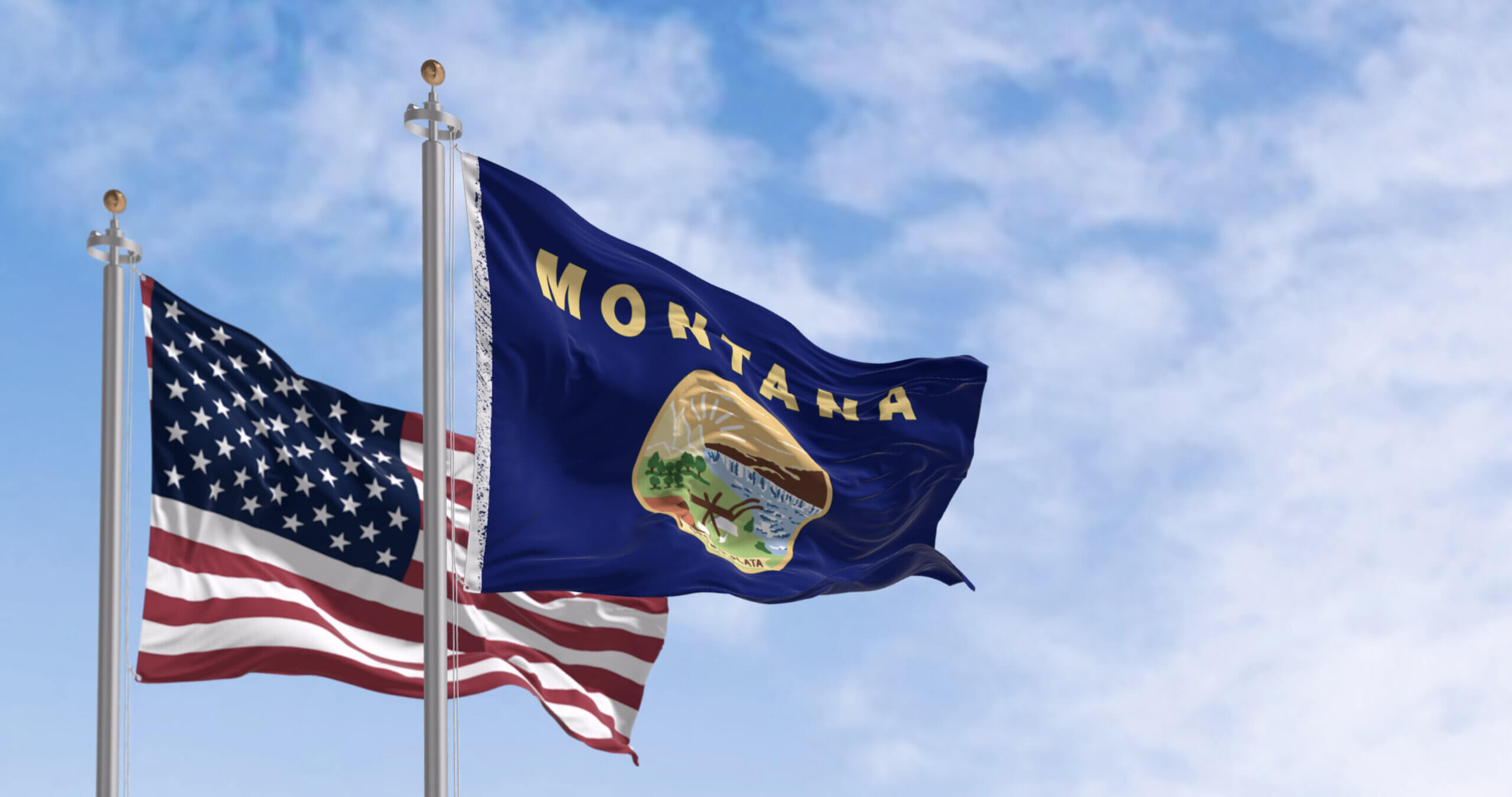 Two flags waving on flagpoles against a blue sky: the U.S. flag with stars and stripes, and the Montana state flag featuring a landscape and "Montana" written in yellow.
