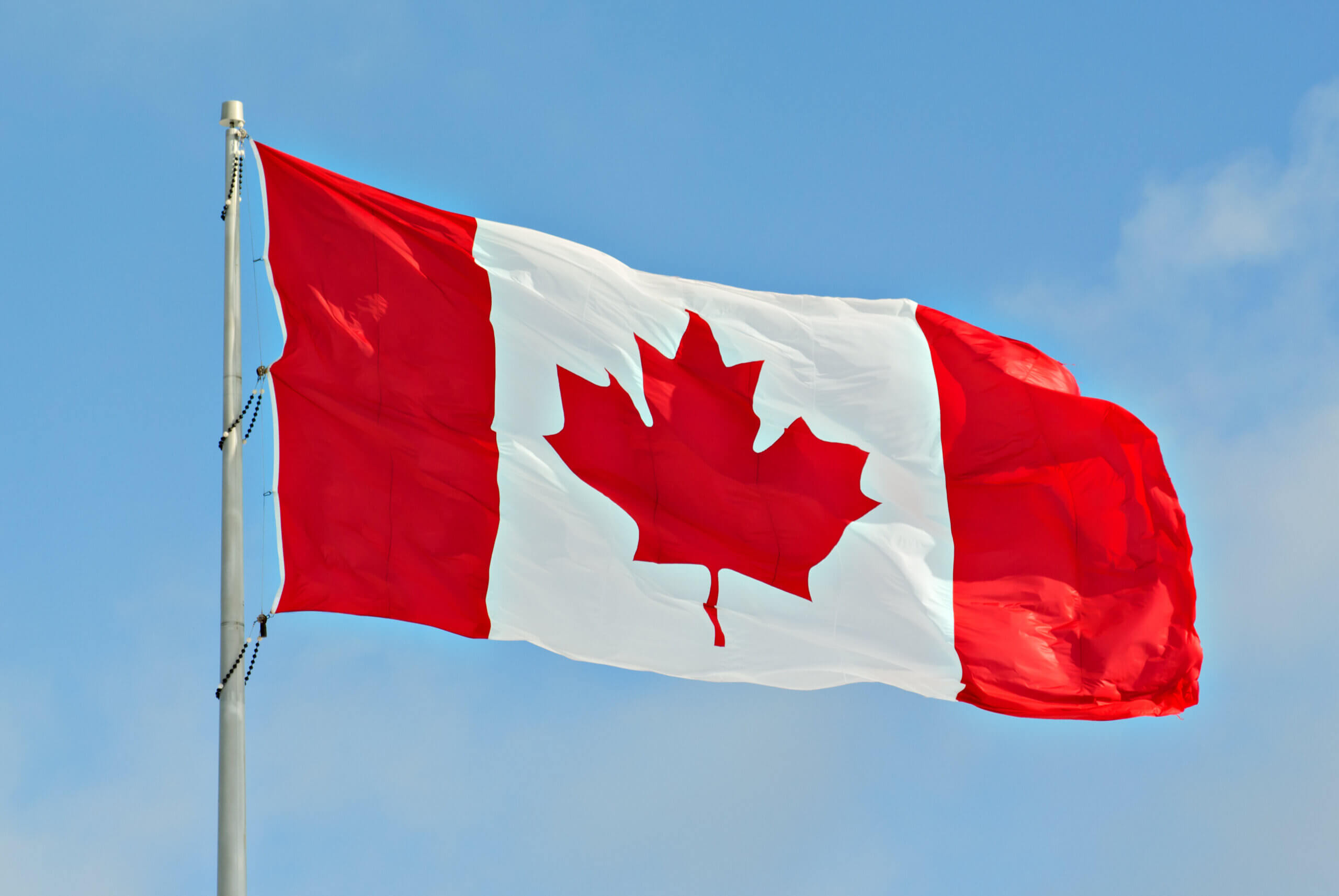 The image shows the flag of Canada waving against a clear blue sky. The flag consists of two vertical red bands on each side and a white square in the middle featuring a red maple leaf.