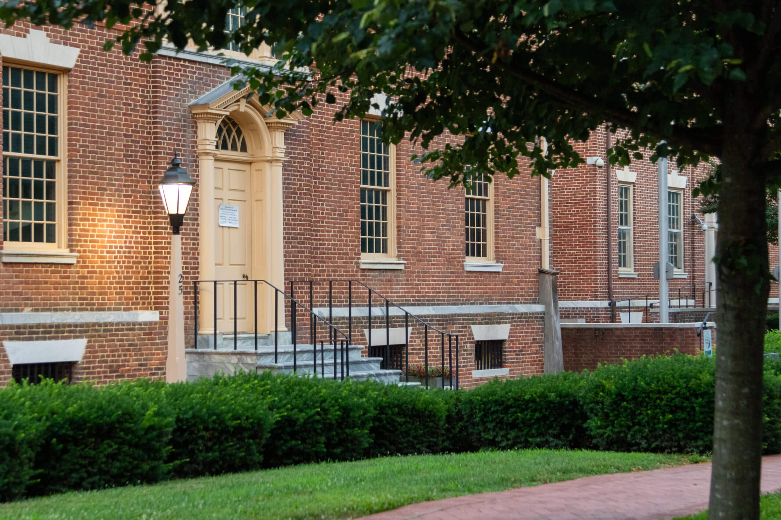 Historic brick building with large windows and a wooden door, surrounded by green hedges and trees. A lantern is mounted on the wall near the entrance. The sidewalk is paved with red bricks, leading to a peaceful, well-maintained lawn.