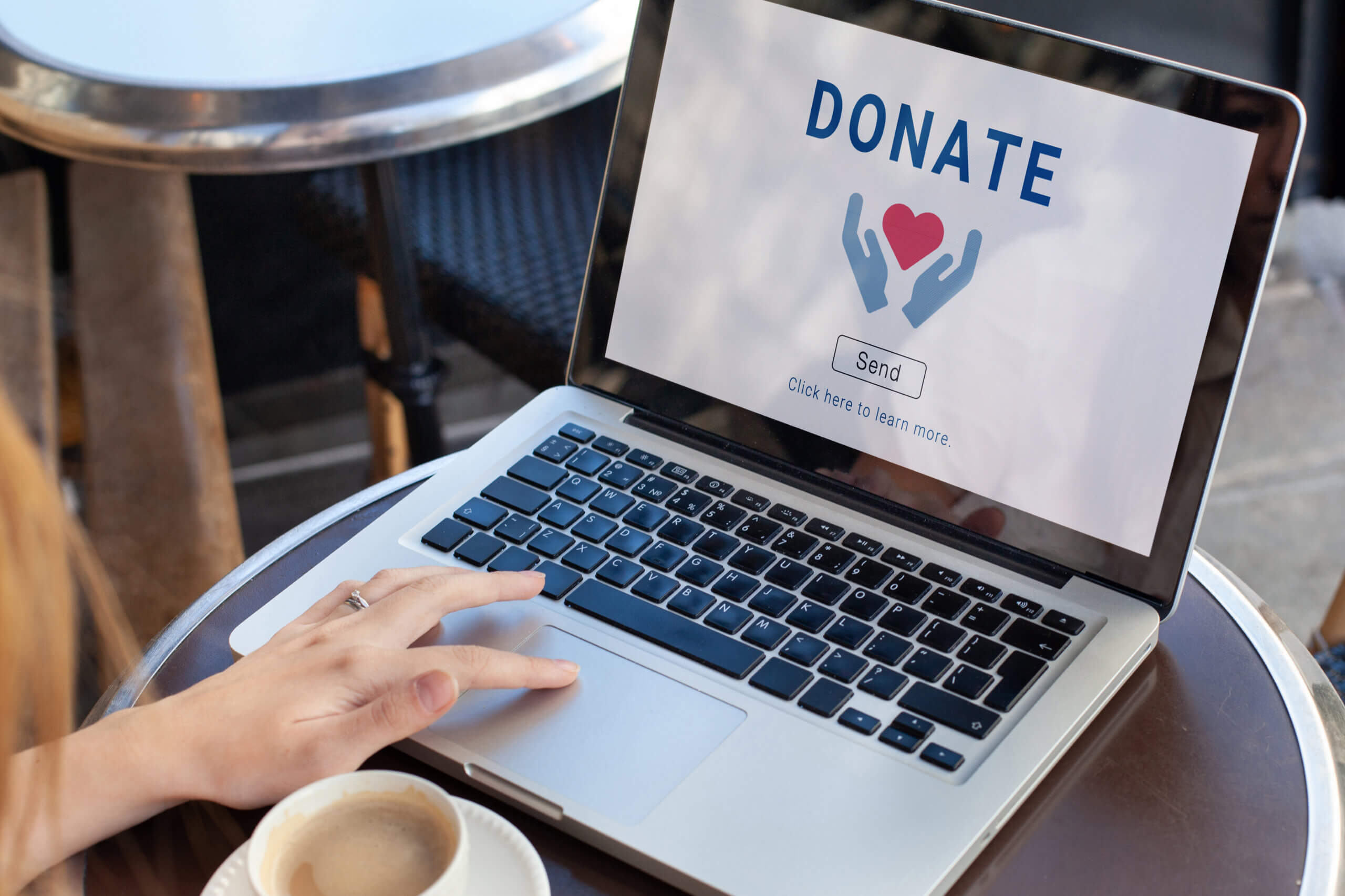 A person is sitting at a table with a cup of coffee, using a laptop. The screen displays the word "DONATE" with an icon of two hands holding a heart, above a "Send" button. The background shows outdoor seating.