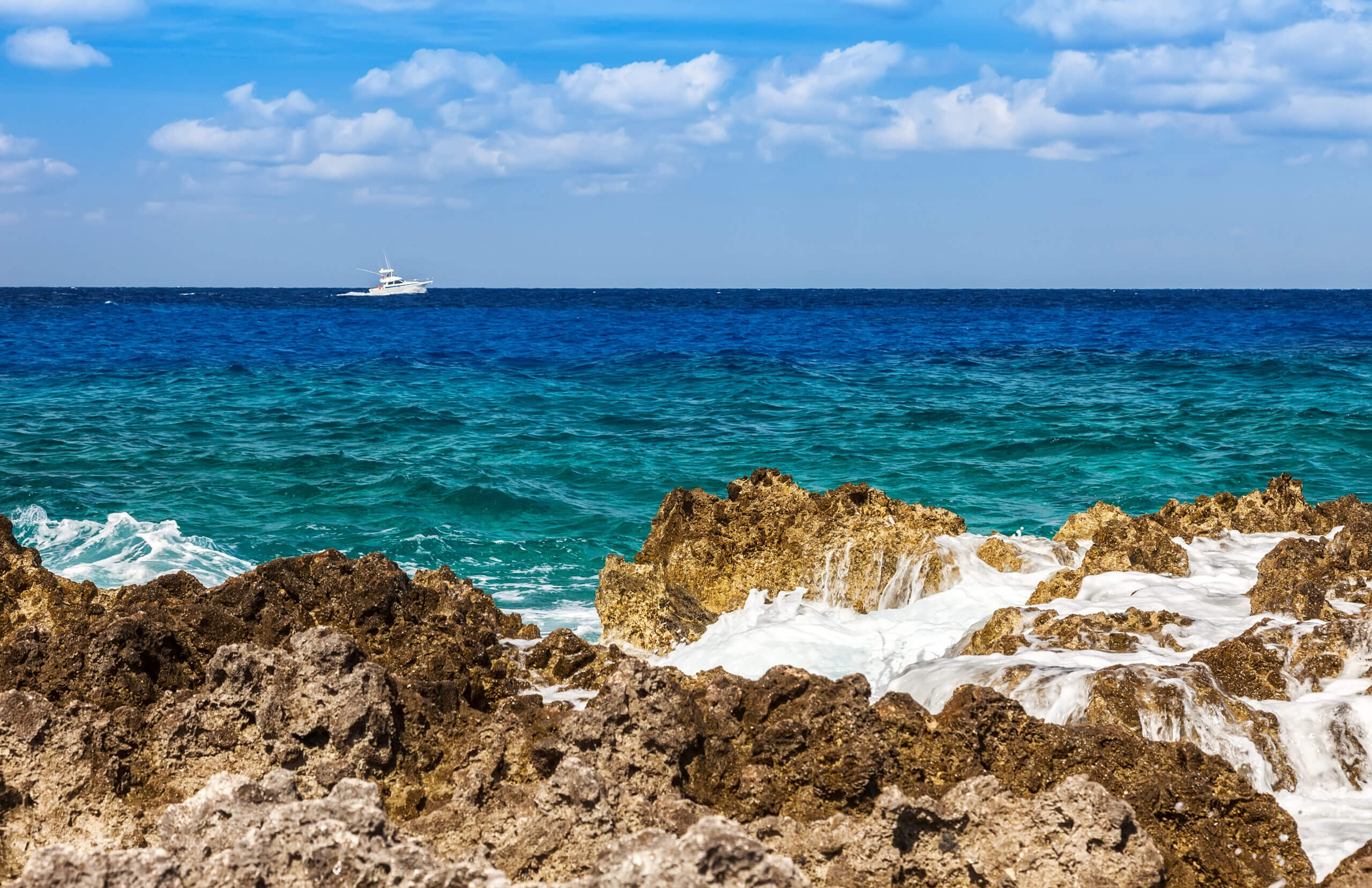 Rocky shoreline with foamy waves crashing against the rocks in the foreground. Deep blue ocean stretches to the horizon, with a white boat visible in the distance under a partly cloudy sky.