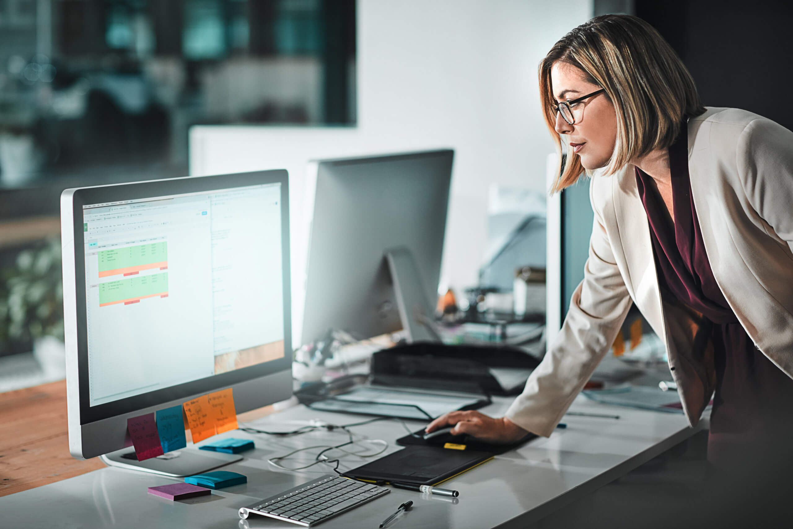 A woman with glasses and short hair leans over a desk, using a computer mouse. She is focused on a large monitor displaying charts and graphs. The desk has colorful sticky notes and office supplies. Other computer monitors are in the background.