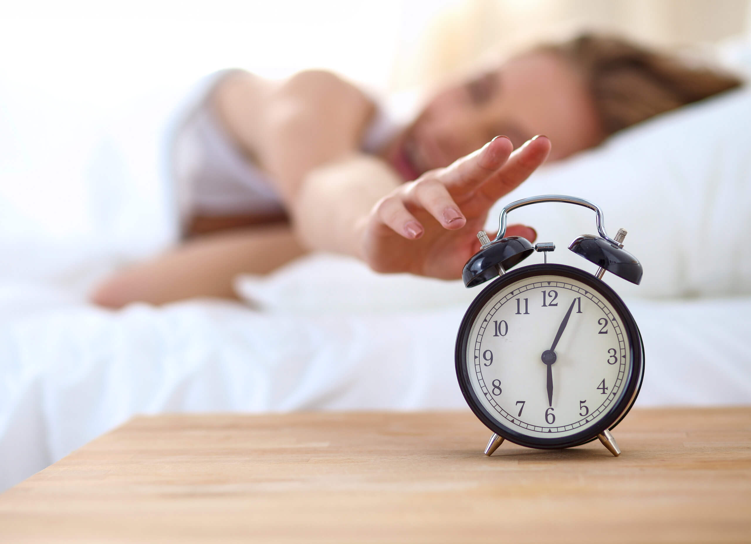 A blurry person reaches out on a bed toward a ringing vintage alarm clock displaying 7:00 on a wooden bedside table.
