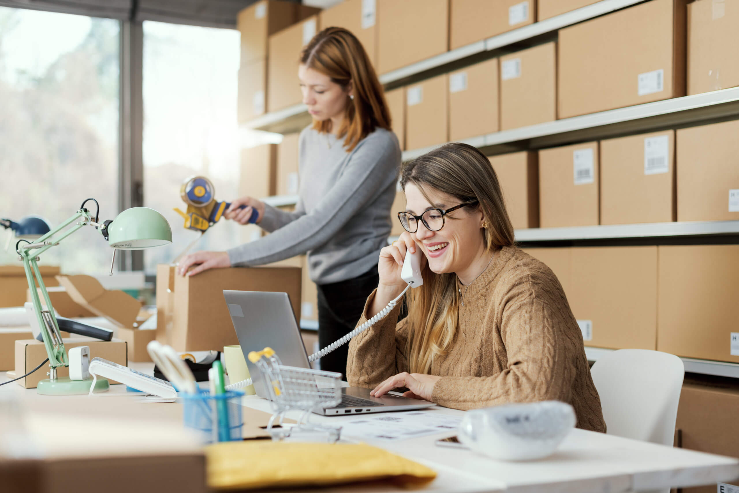 Two women are in a room with shelves of cardboard boxes. One is on the phone at a desk with a laptop, while the other is taping a box. The setting appears to be an office or warehouse.