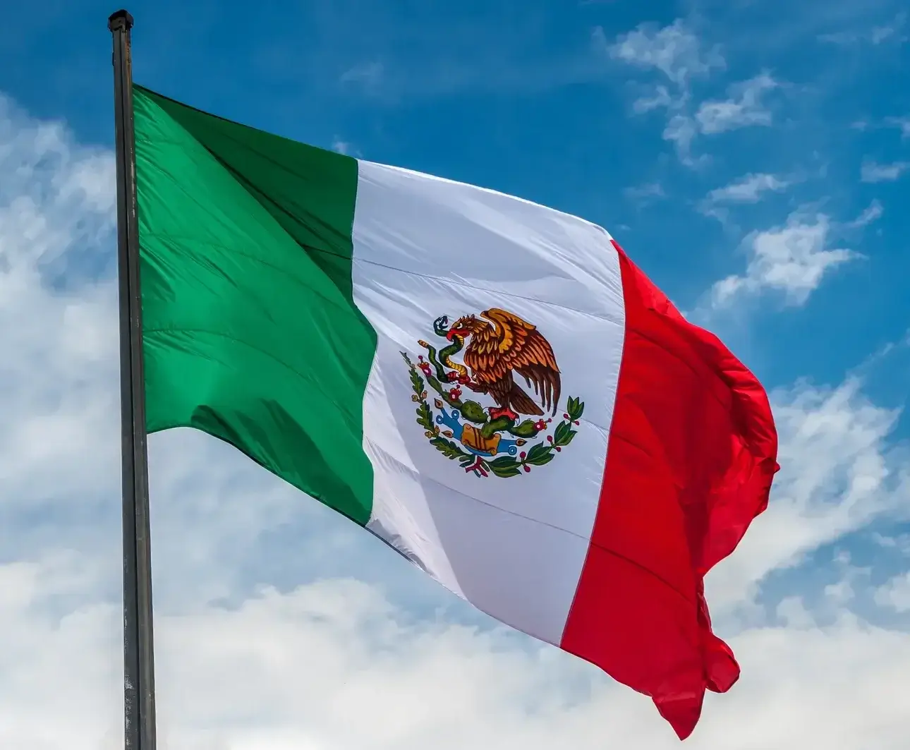 The image shows the Mexican flag waving against a backdrop of a blue sky with scattered clouds. The flag has vertical stripes of green, white, and red, with the national coat of arms featuring an eagle and snake in the center.