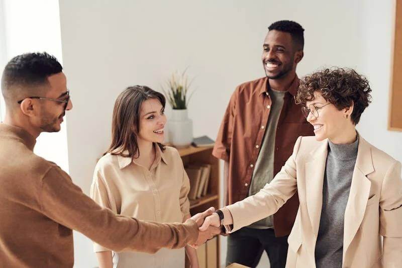Four people in a bright office setting are engaged in a friendly conversation. Two individuals are shaking hands, and they all appear to be smiling. A plant and shelves are visible in the background.