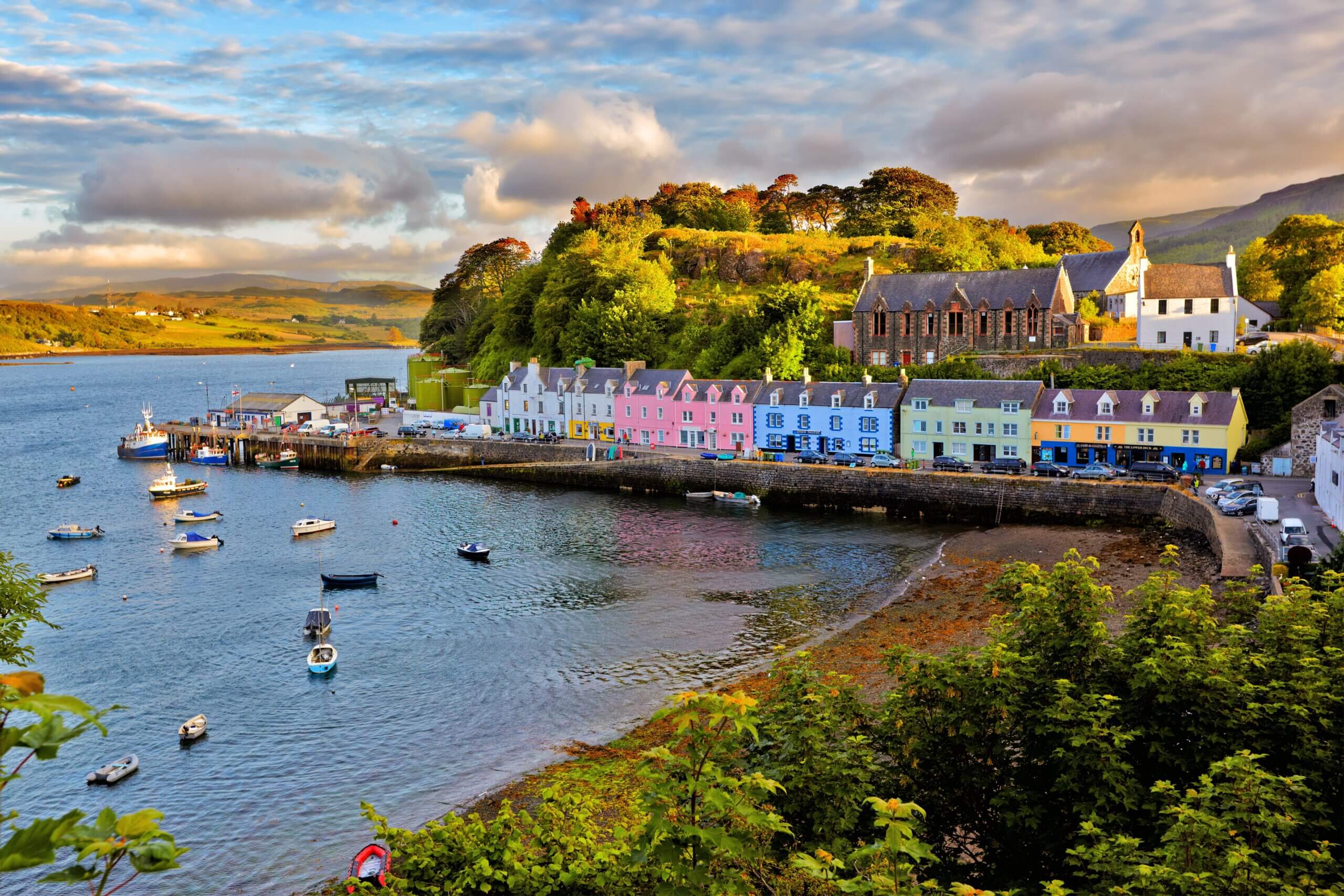 Colorful buildings line the waterfront of a small harbor at sunset, with boats moored in the calm water. Lush greenery and trees are on the hill behind, and the sky is partly cloudy with warm sunlight illuminating the scene.