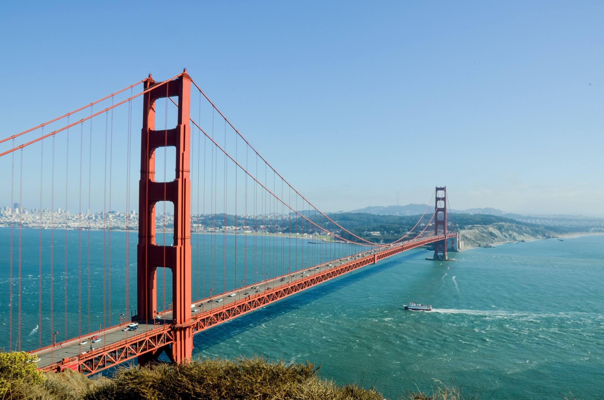 The image shows the Golden Gate Bridge spanning across the blue water, with hills in the background under a clear sky. A boat leaves a white trail in the water below the bridge.