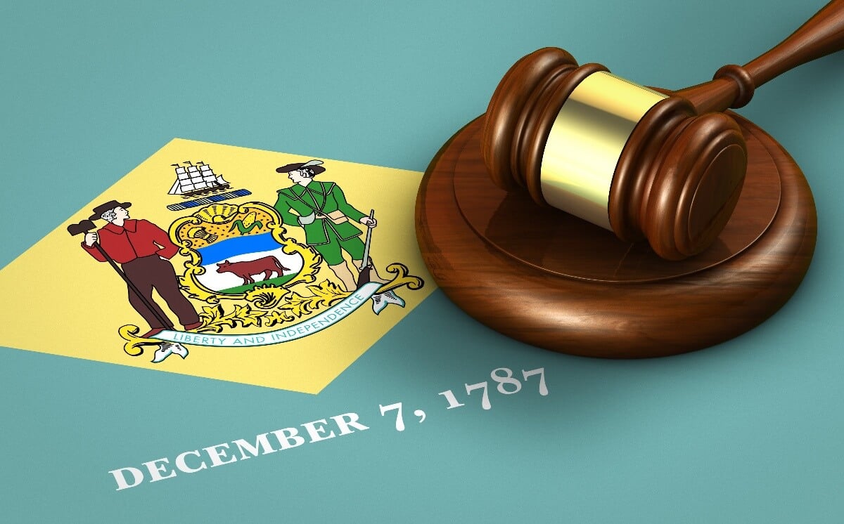 A gavel rests on a surface next to the Delaware state flag, featuring a farmer, a soldier, a ship, corn, wheat, an ox, and the motto "Liberty and Independence." Below, the date "December 7, 1787" is displayed.