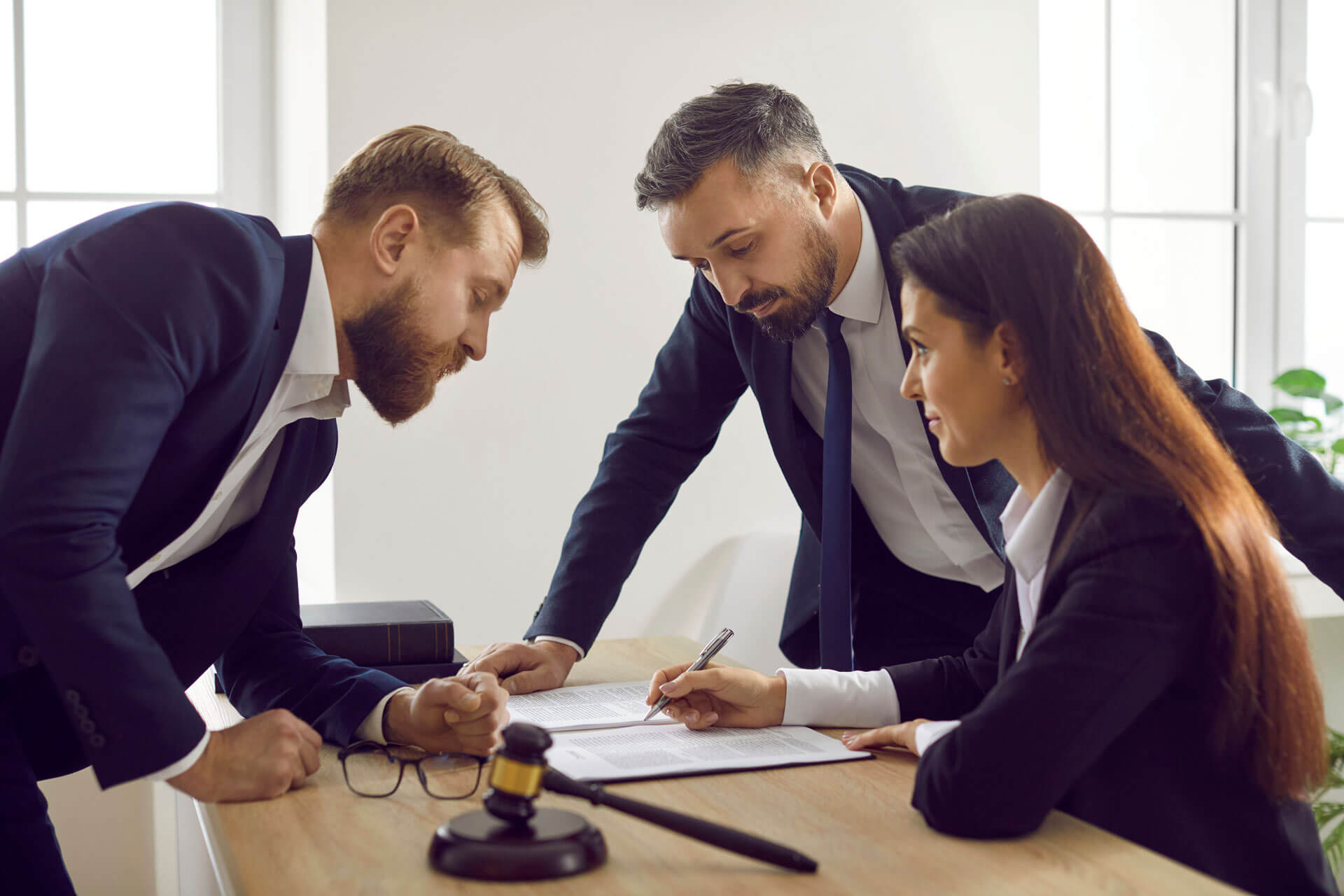 Three people in business attire are at a table in an office setting, discussing documents. One woman is seated, writing on a paper, while two men stand over the table, engaged in conversation. A gavel is visible on the table.