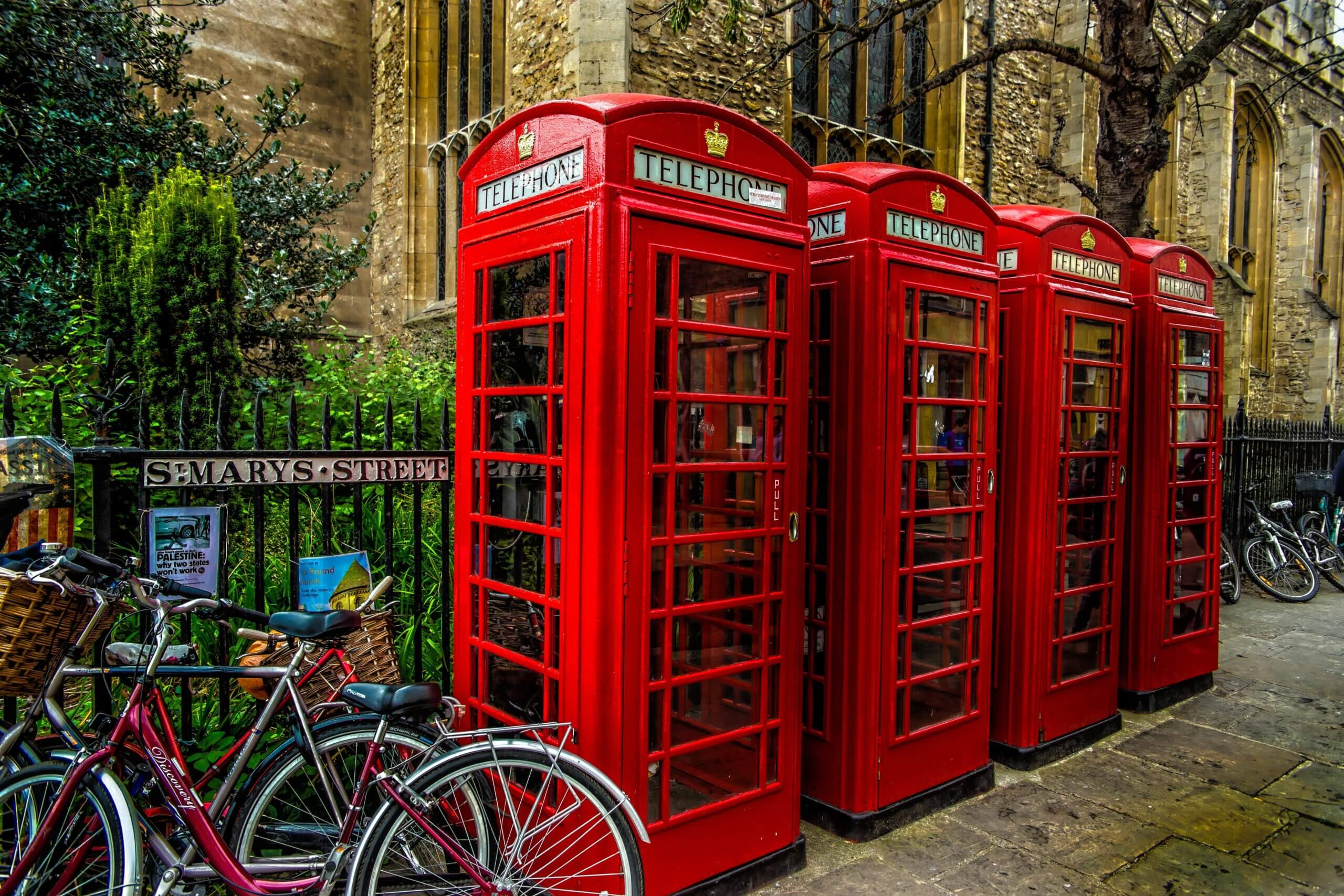 Four classic red British telephone booths stand in a row near a stone building. Two bicycles are parked in front of them. A sign reads "St. Mary's Street," and there are trees and greenery around.