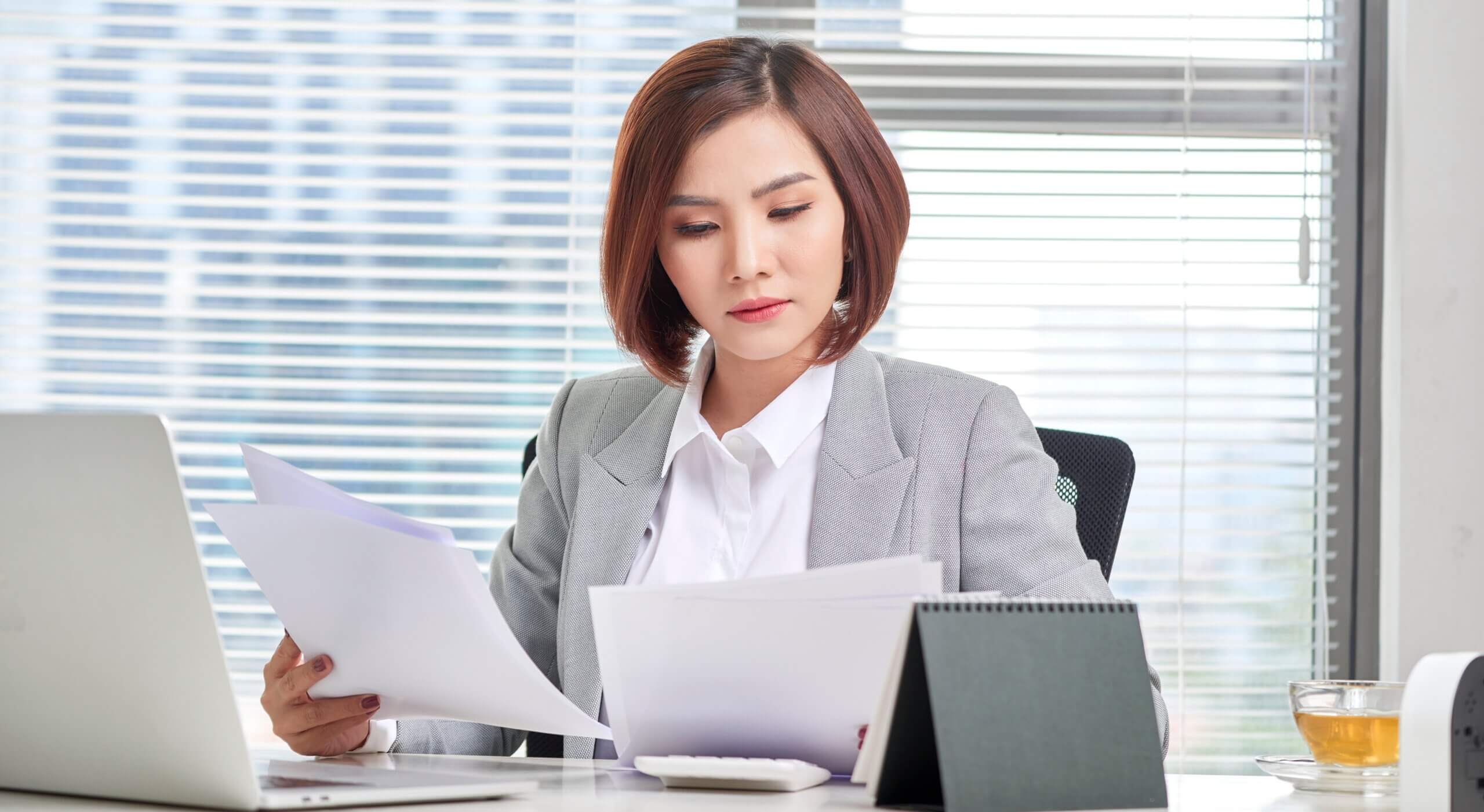 A woman with short hair in a gray suit is seated at a desk, reviewing documents. She appears focused. In front of her are a laptop, a tablet stand, and a cup with a drink. Blinds cover a large window behind her.