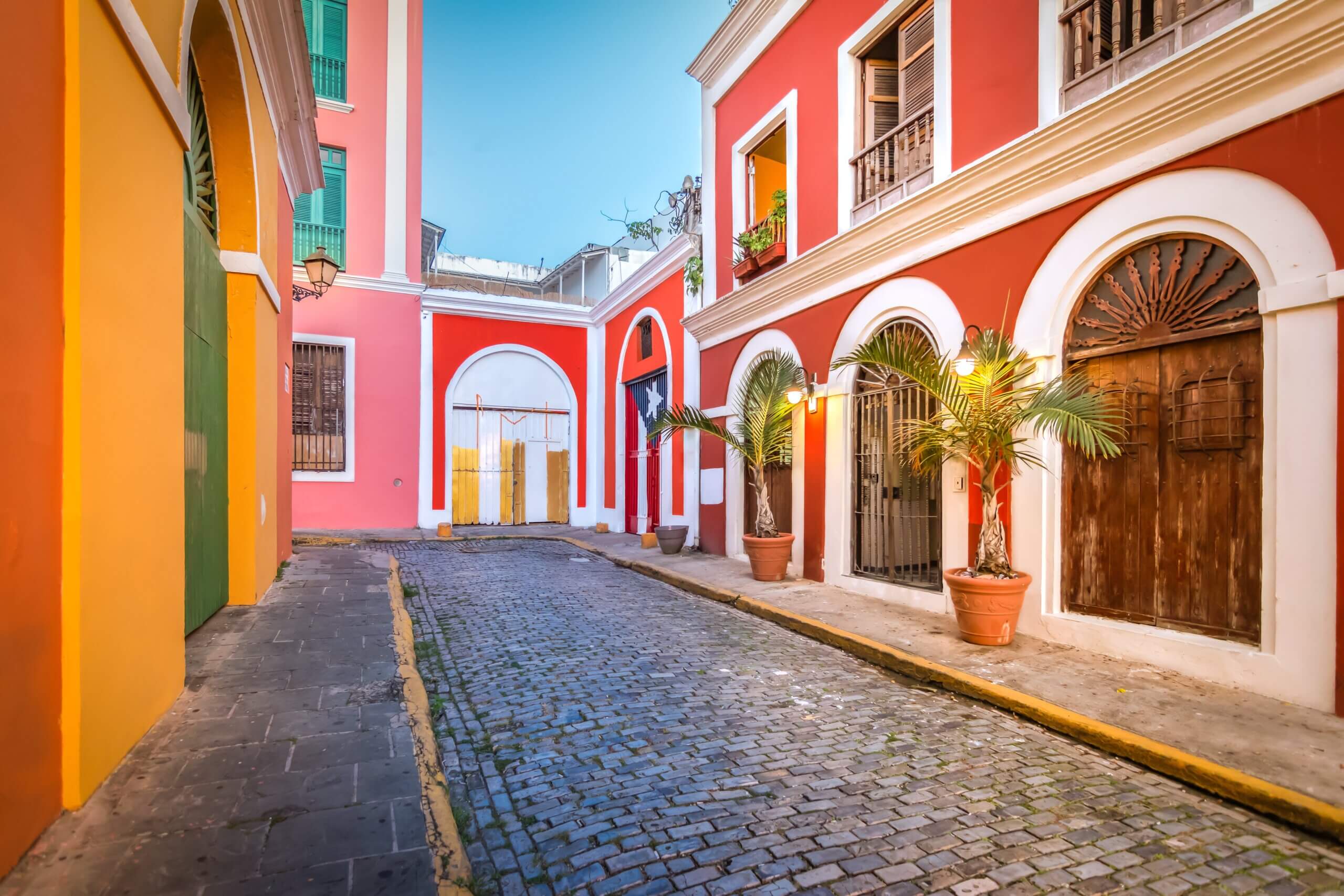 A vibrant street scene with colorful buildings in red, orange, and yellow. The cobblestone path is lined with arches and potted palm plants. Warm lighting from lamps creates a cozy atmosphere under a clear sky.