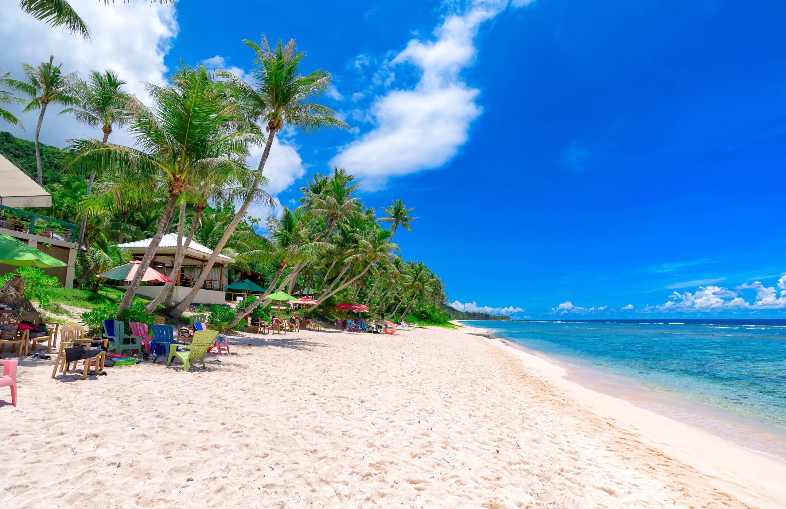 A tropical beach scene with white sand and clear blue water. Palm trees line the shore, alongside colorful beach chairs and umbrellas. A few people relax in the shade, with a bright blue sky overhead.