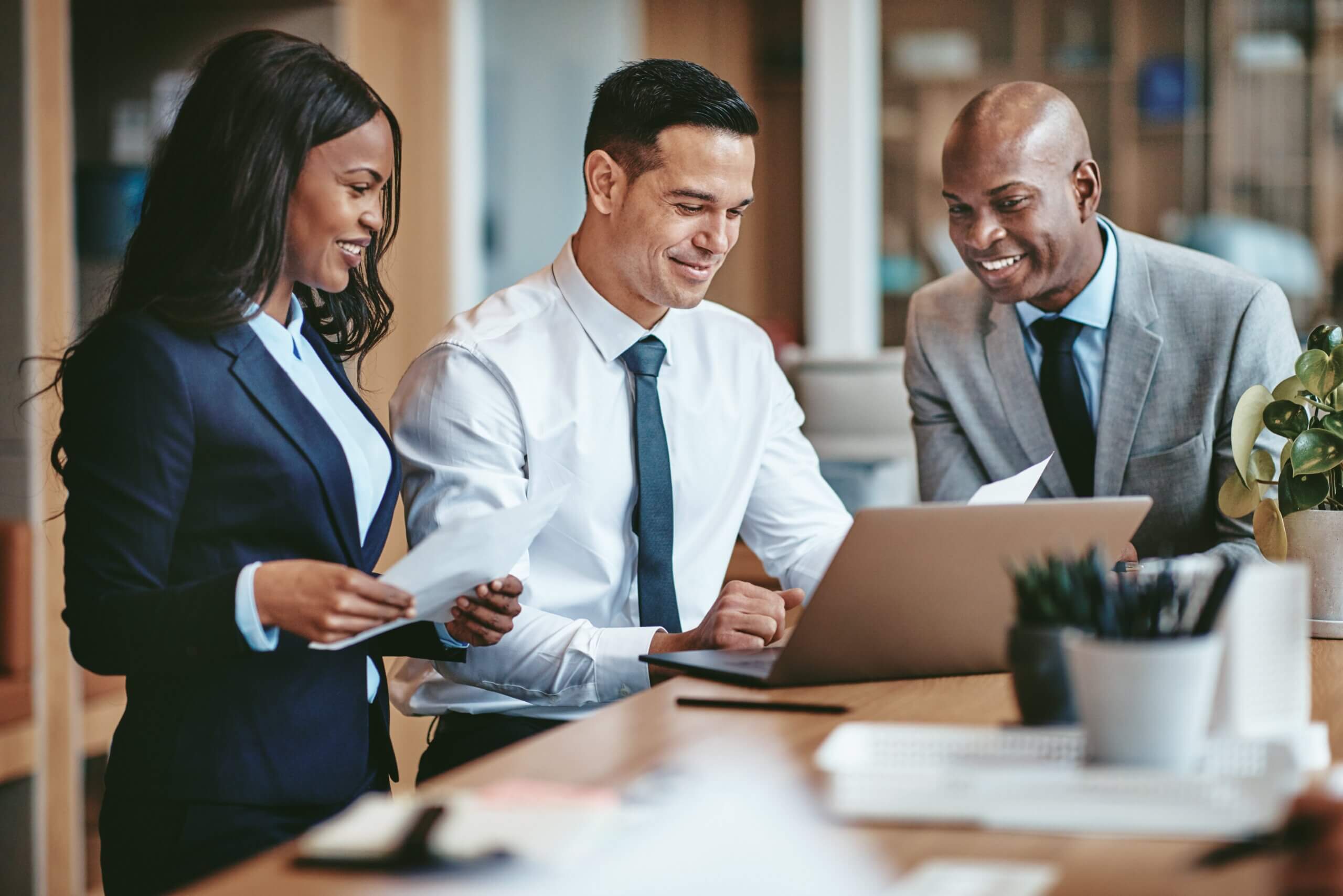 Three business professionals, two men and one woman, are gathered around a desk in an office setting, smiling and looking at documents and a laptop. They are dressed in formal suits and appear engaged in a discussion.