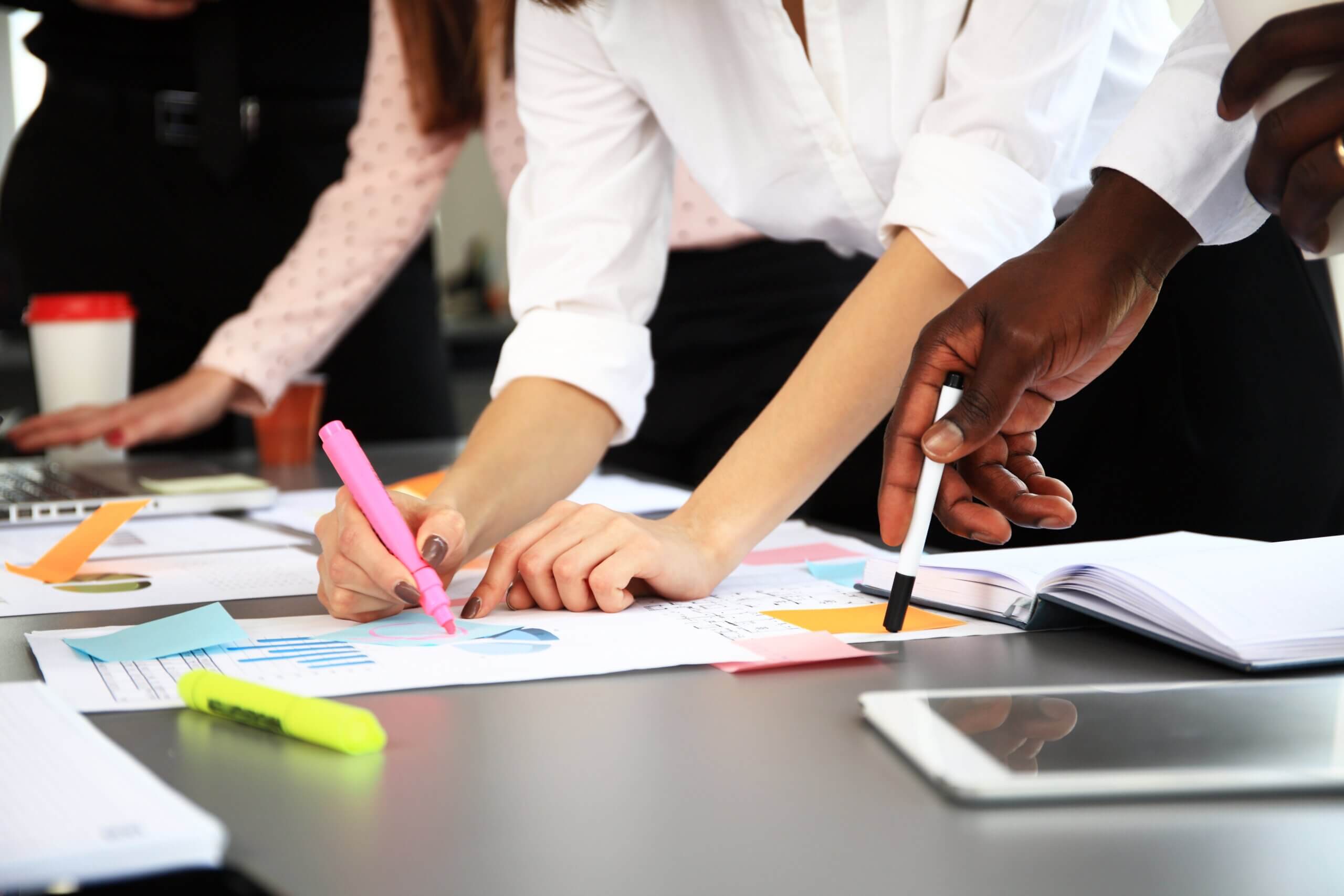 People collaborate around a table covered with documents, charts, and a tablet. One person uses a pink highlighter, while another points with a pen. Highlighters, a cup, and notebooks are visible, suggesting a meeting or brainstorming session.