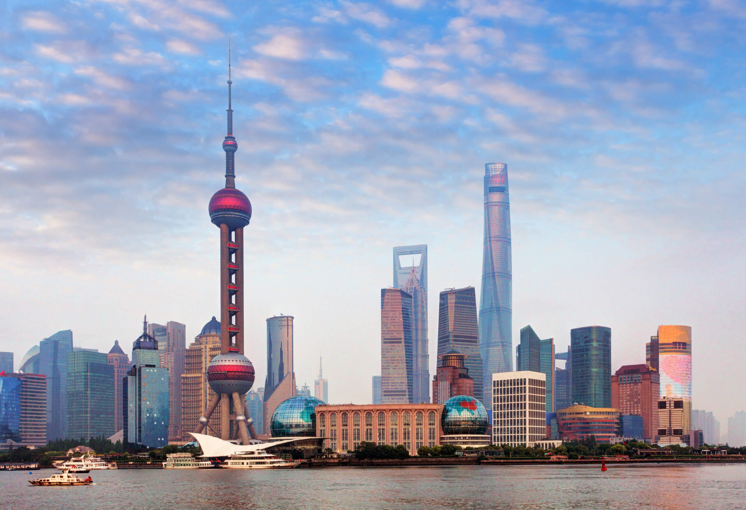 Shanghai skyline featuring the Oriental Pearl Tower and modern skyscrapers under a cloudy sky, with boats on the Huangpu River in the foreground.