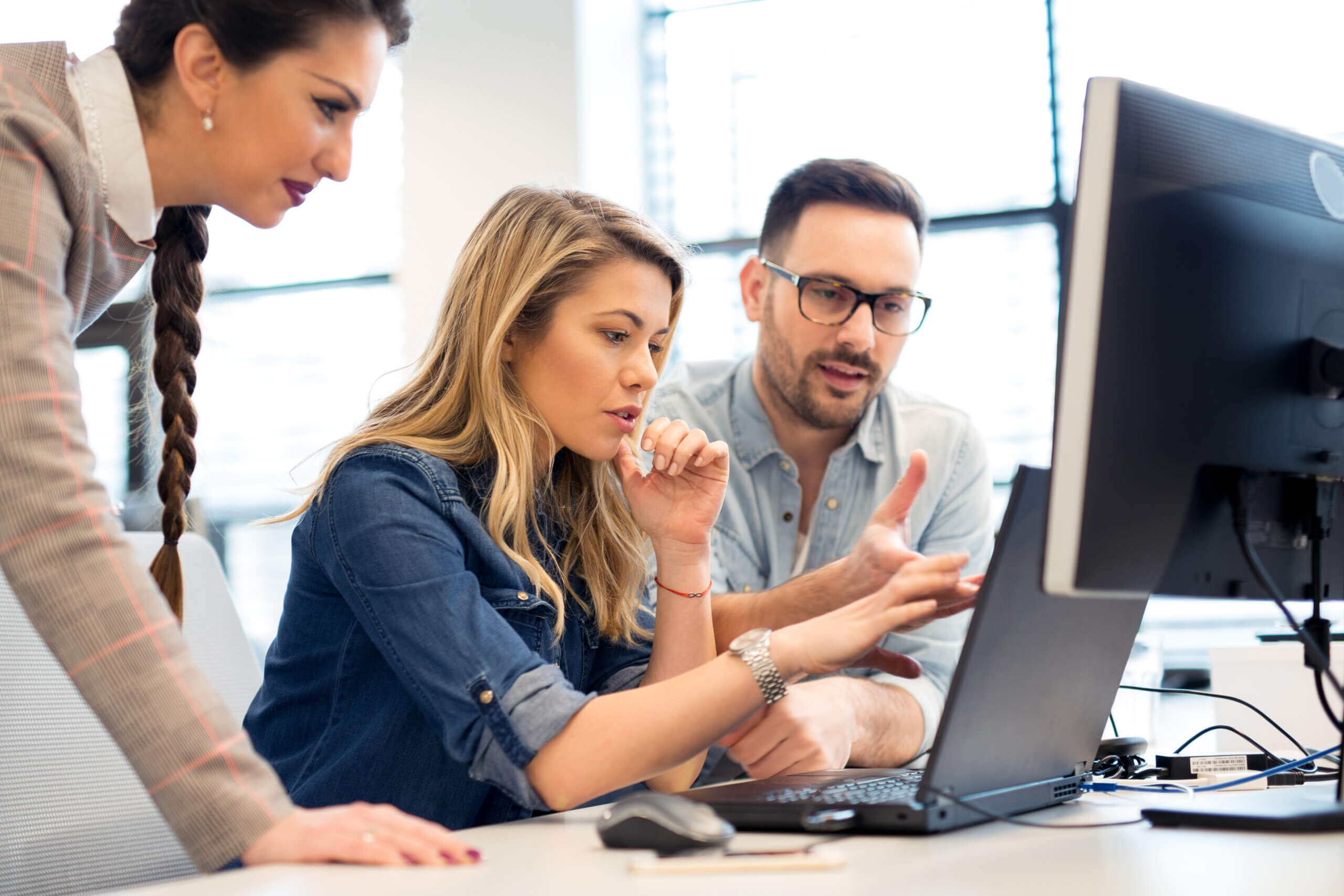 Three people collaborating at a desk, focused on a computer screen. The woman in the middle points at the monitor, while the man gestures and the other woman observes intently. The room is well-lit with large windows in the background.