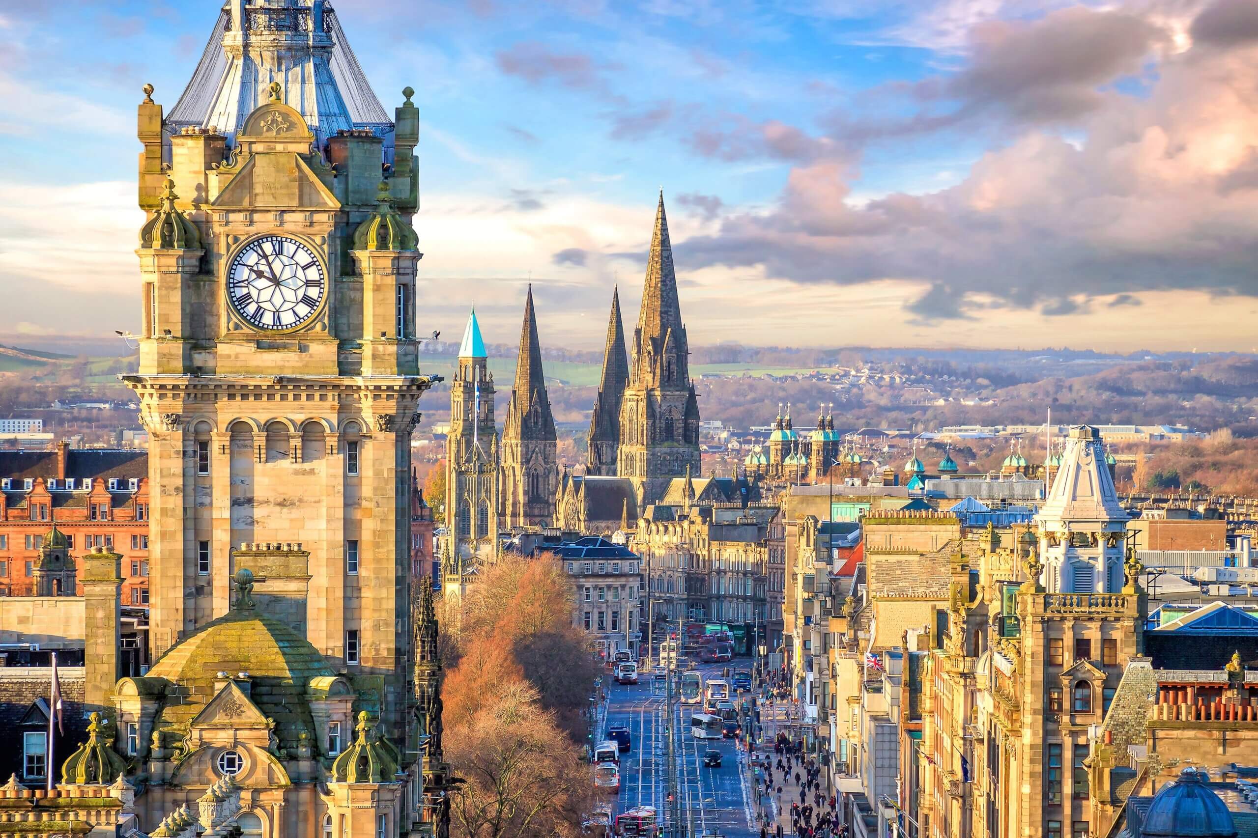 Aerial view of a historic cityscape featuring a prominent clock tower with two clocks, surrounded by ornate buildings and several tall spires. Leafless trees line the streets, and a partly cloudy sky is in the background.