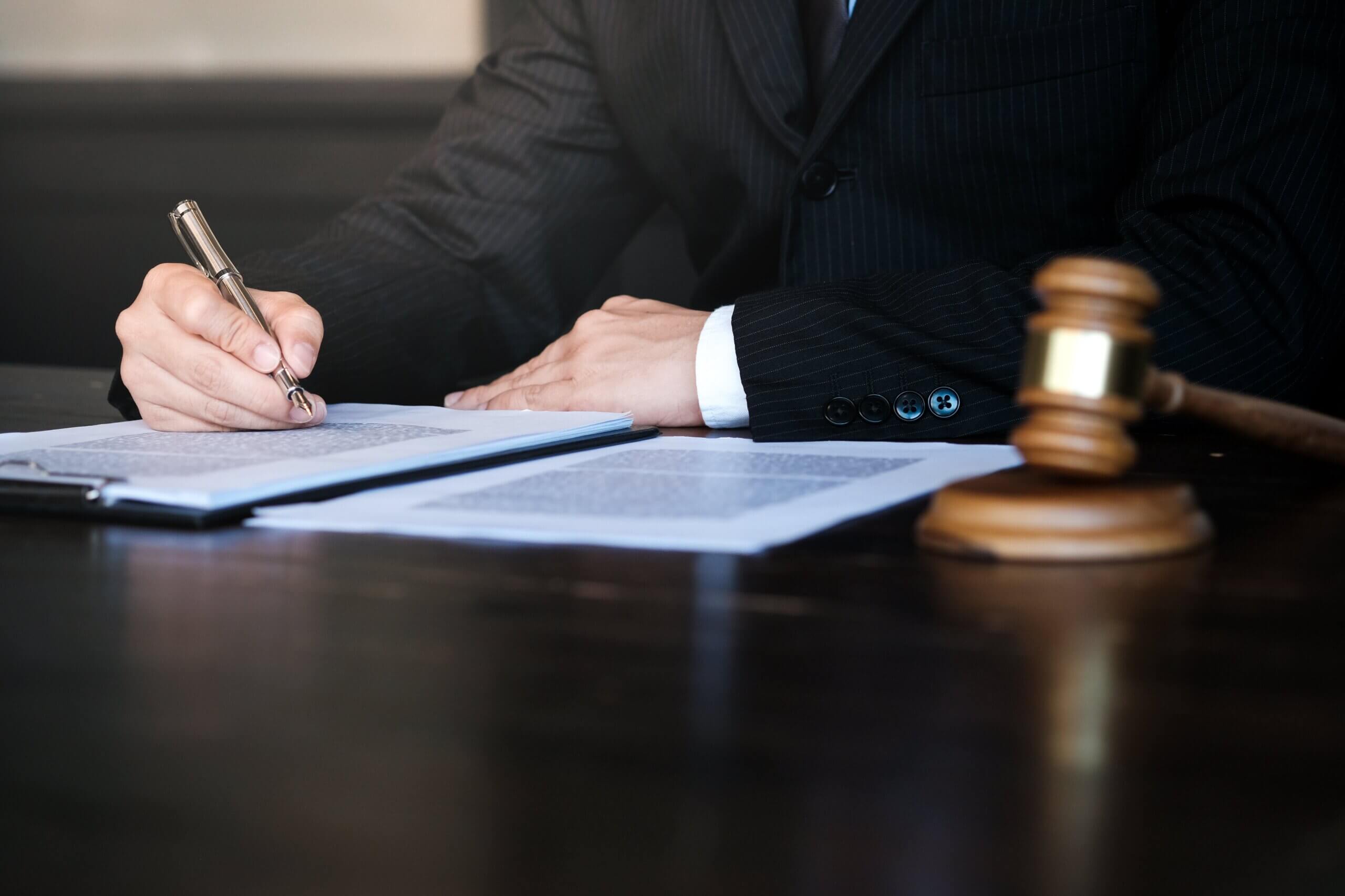 A person in a suit is writing on a document at a desk. A wooden gavel is nearby, symbolizing law or justice. The focus is on the hand holding a pen, with papers spread out on the dark wooden surface.