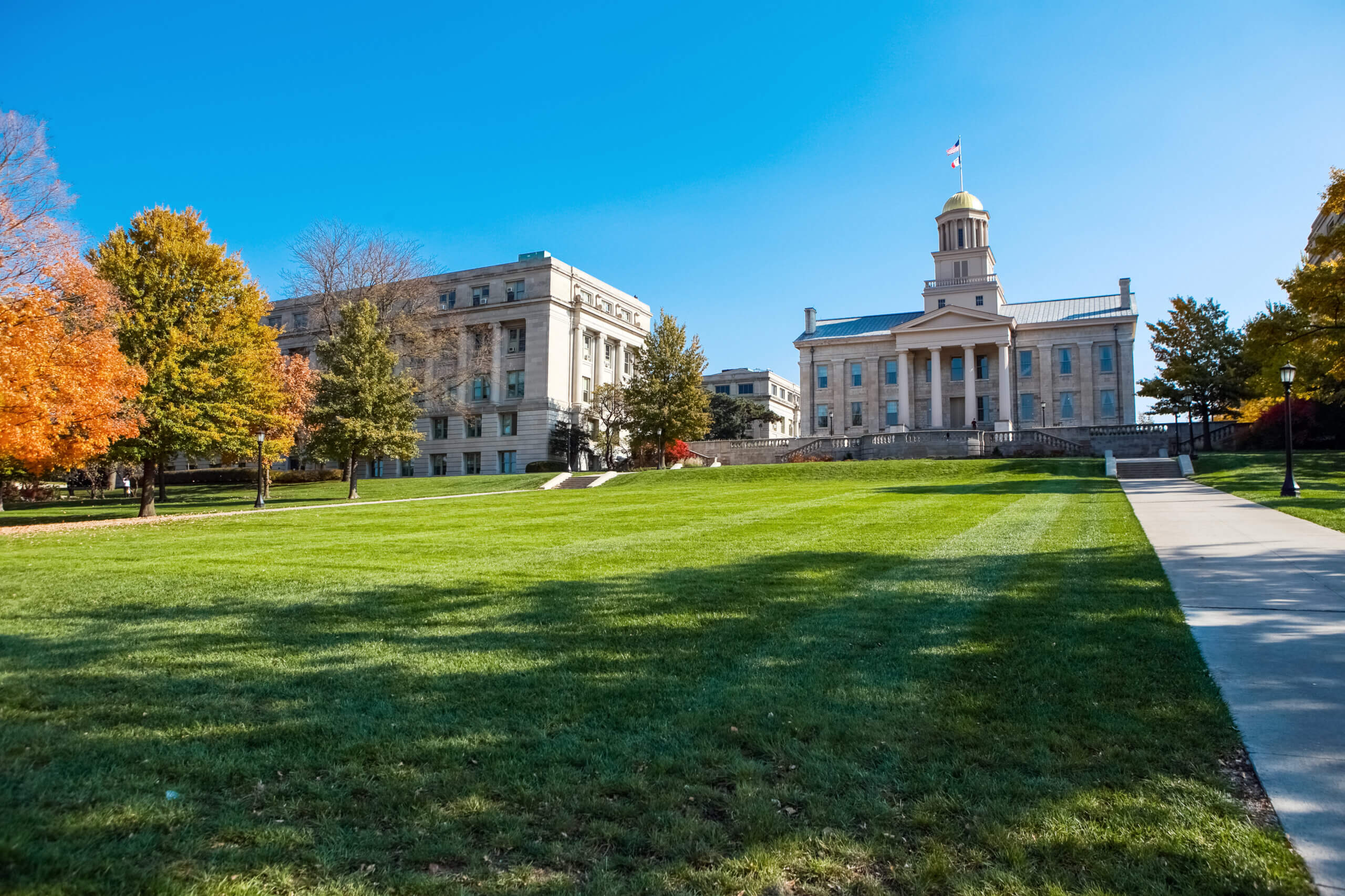 A historic building with a tall, columned entrance and a dome, surrounded by trees with autumn foliage. A well-manicured lawn leads up to the structure, and a clear blue sky is overhead.