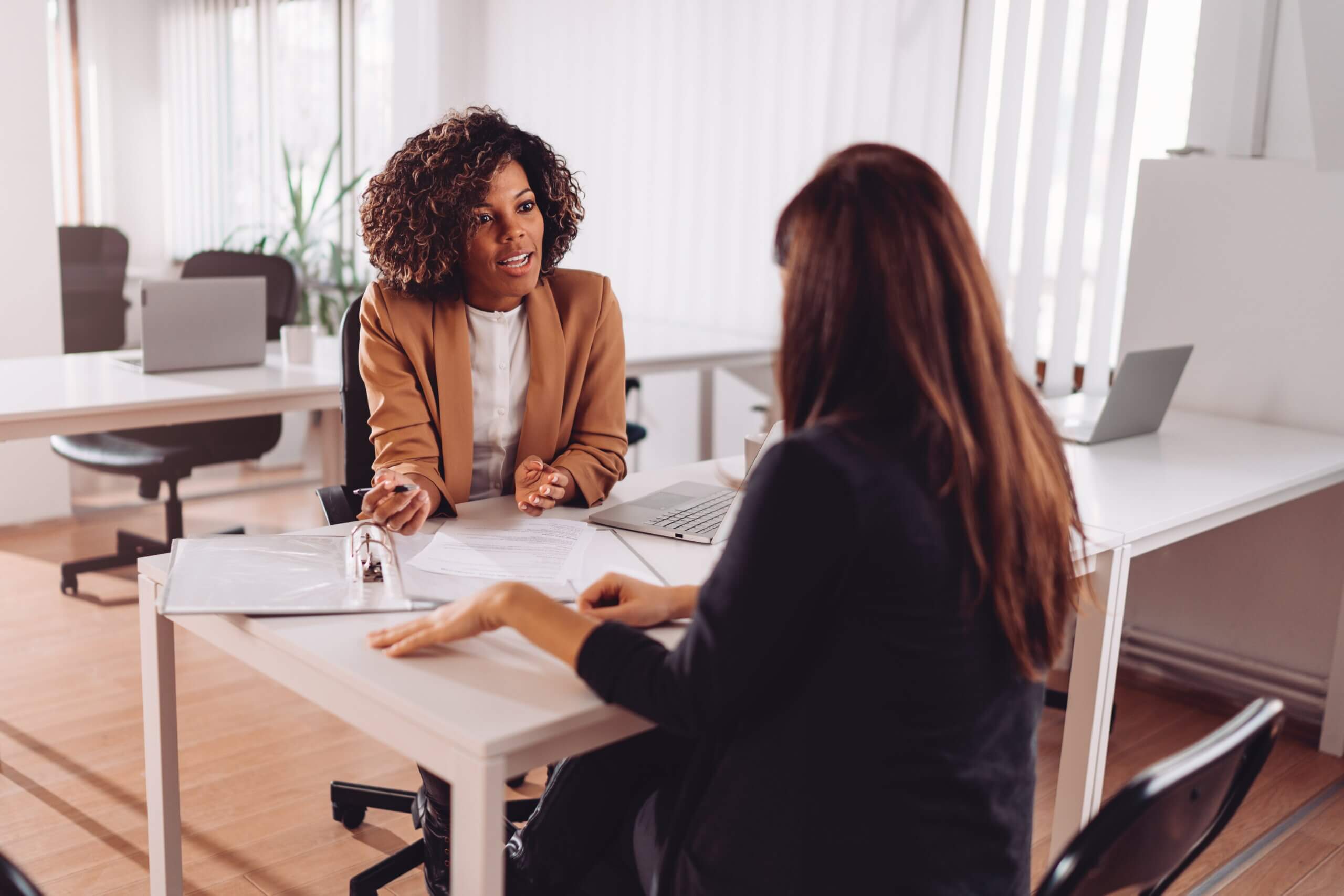 Two women sitting across from each other at a table in an office setting. One woman, wearing a brown blazer, is speaking and gesturing with her hands. Papers and a laptop are on the table. The other woman listens attentively.