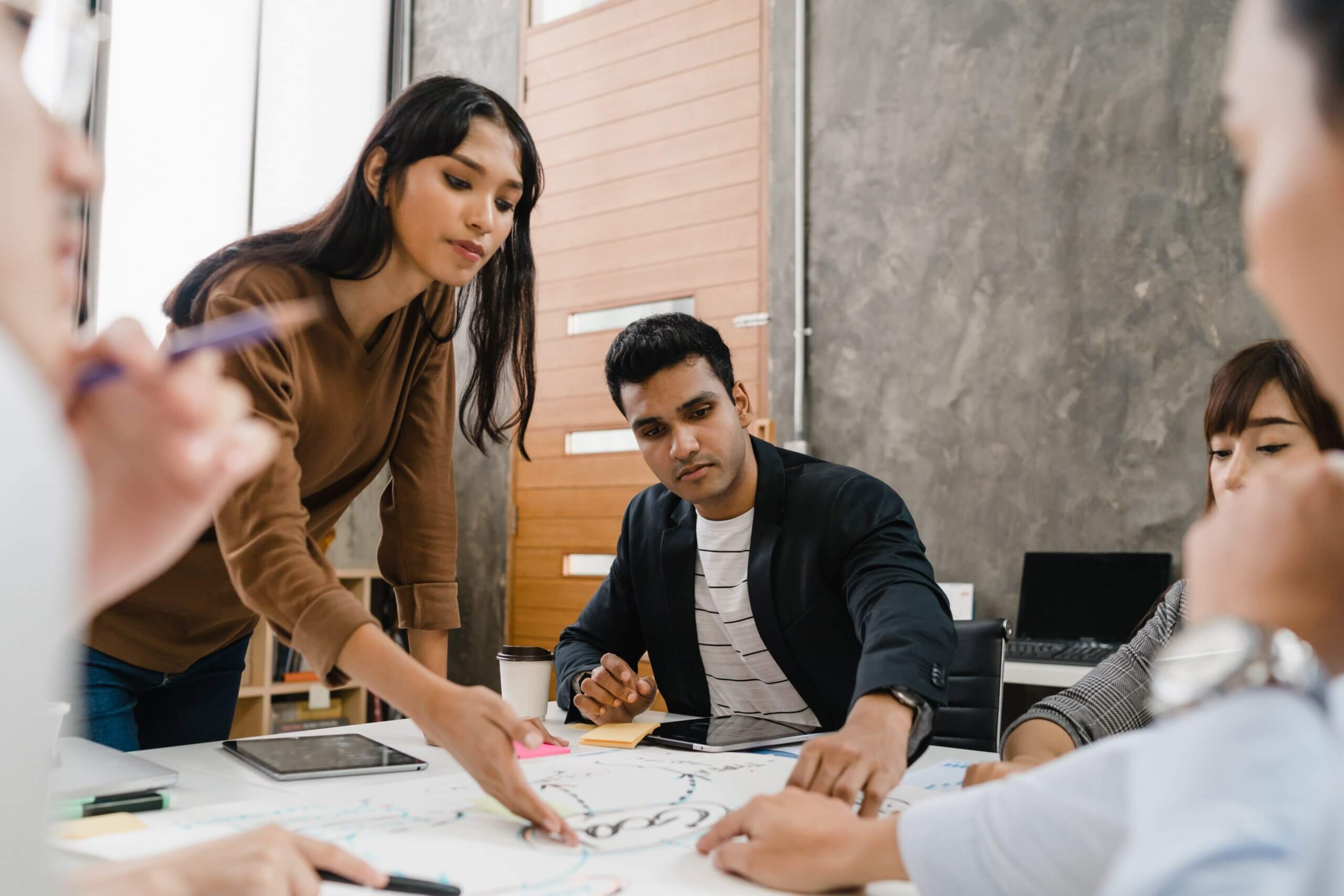 A diverse group of people gathers around a table in a modern office setting. They are engaged in a discussion, with charts and papers spread out in front of them. One woman stands, gesturing towards the documents, while others listen attentively.