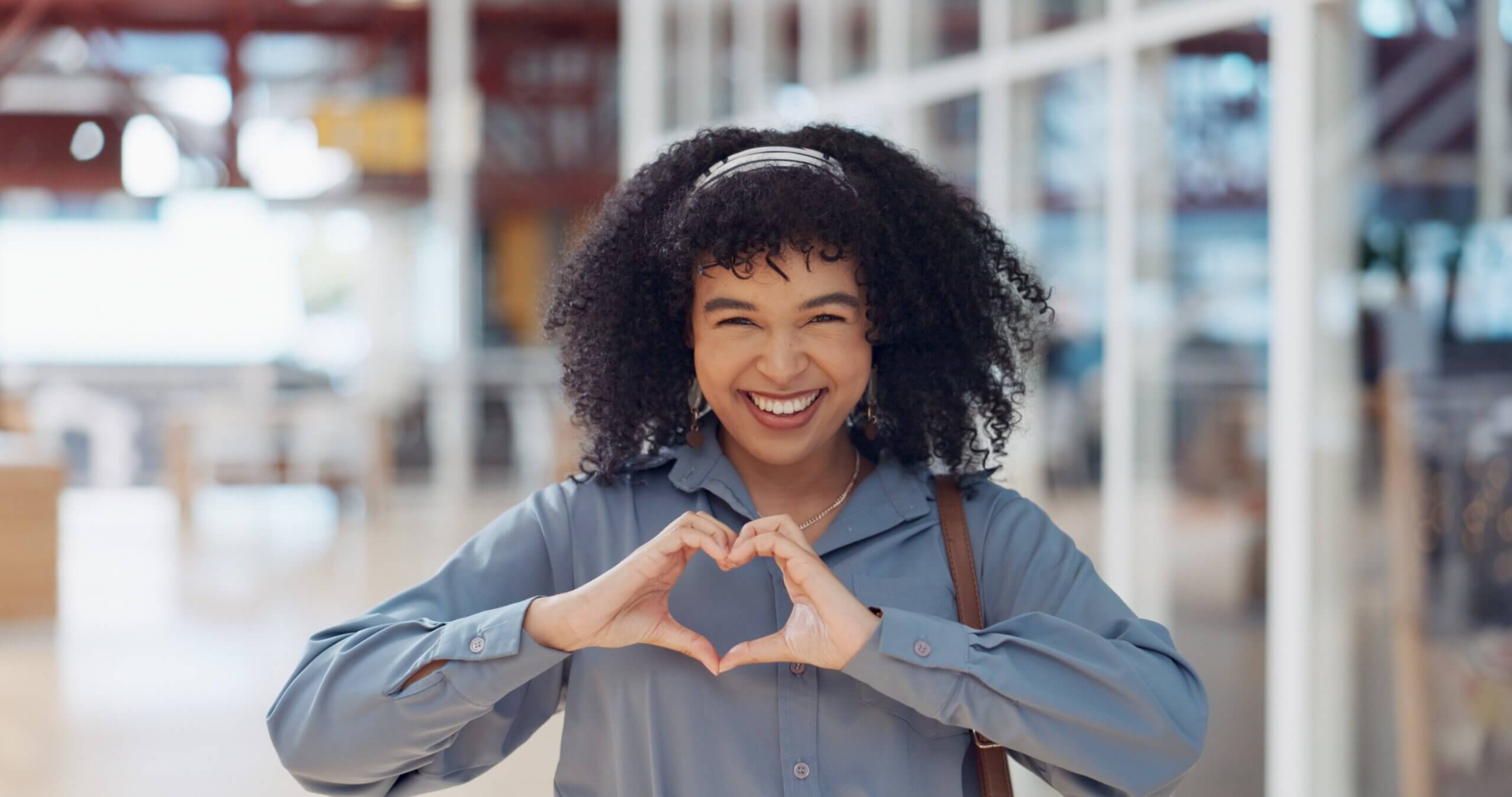 A person with curly hair smiles warmly, making a heart shape with their hands. They are wearing a gray shirt, and the background features a bright, modern interior with glass walls.
