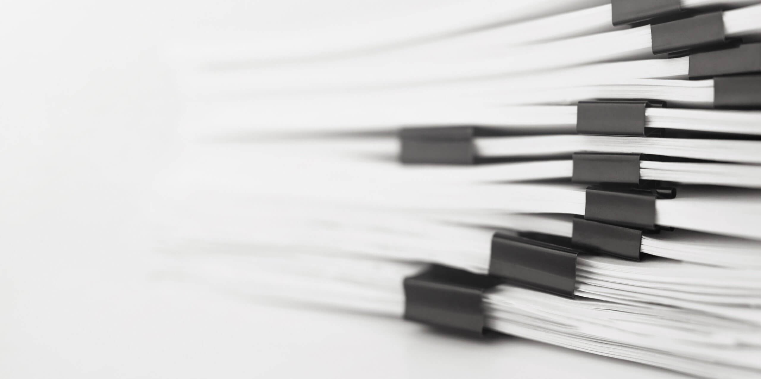 A close-up of several stacks of white papers bound together with black binder clips, placed horizontally. The image background is soft and out of focus, emphasizing the organization of the documents.