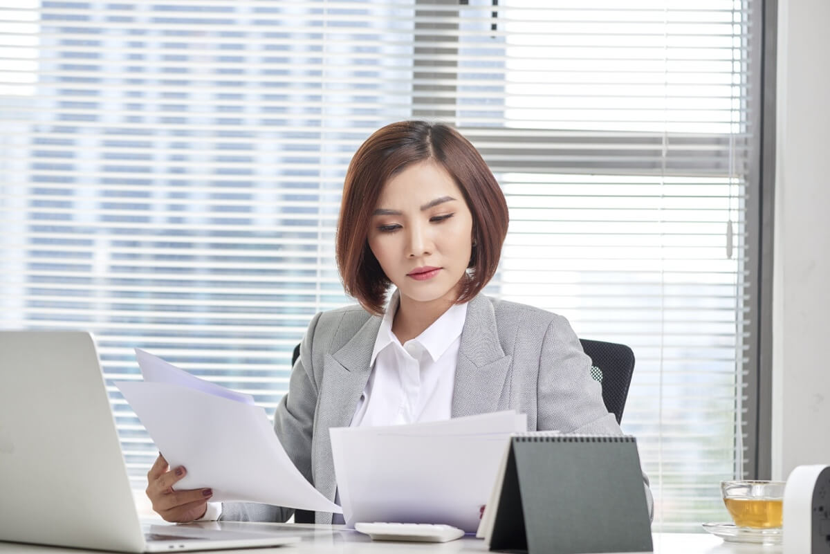 A woman in a gray suit is sitting at a desk, reviewing documents. She has short brown hair and appears focused. There's a laptop, a calendar, and a cup of tea on the desk. Blinds partially cover a window with a city view in the background.