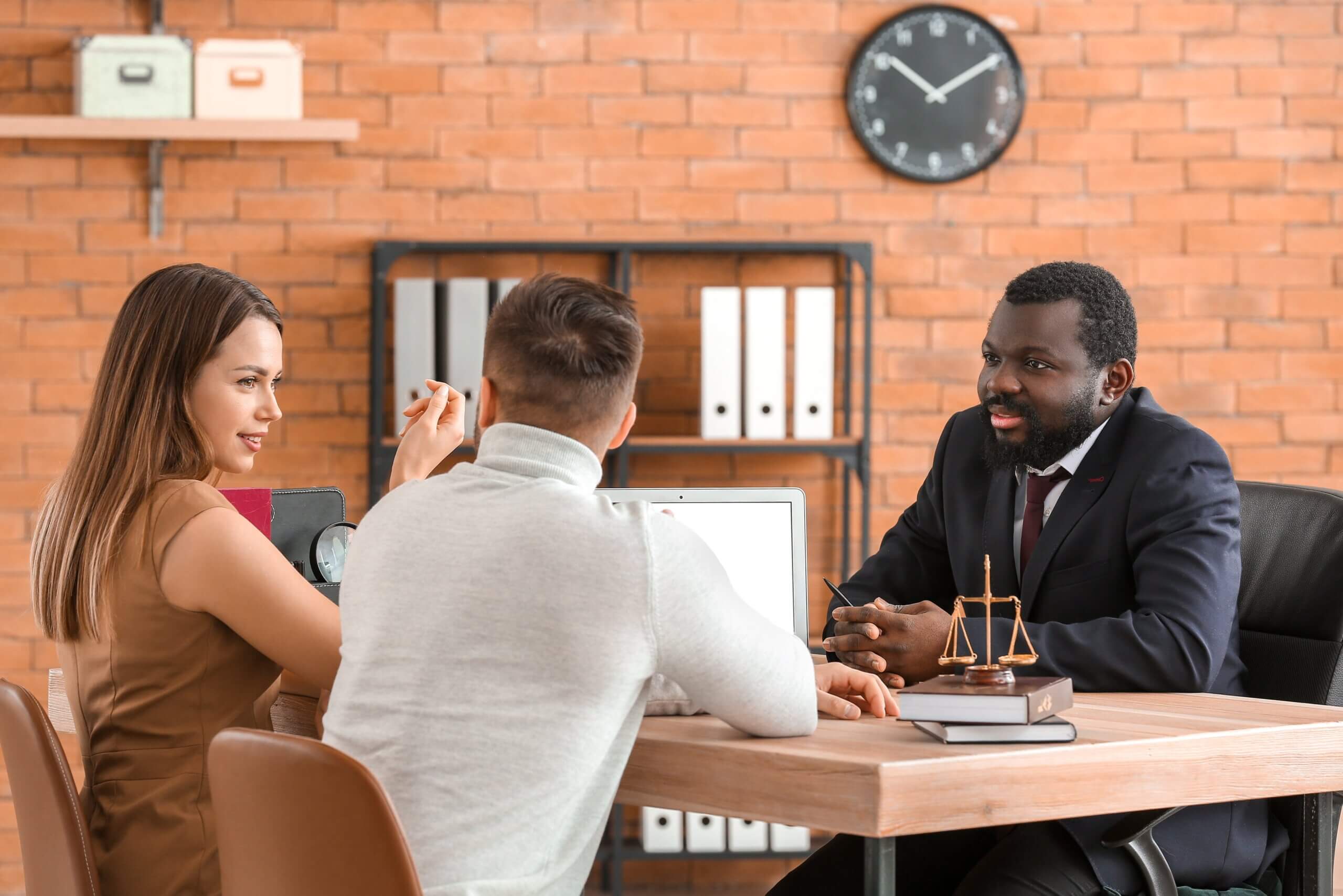 Three people having a discussion in an office. A woman and a man sit on one side of a table, facing another man wearing a suit. The table holds a laptop, books, and scales. A clock and shelves with binders are in the background.